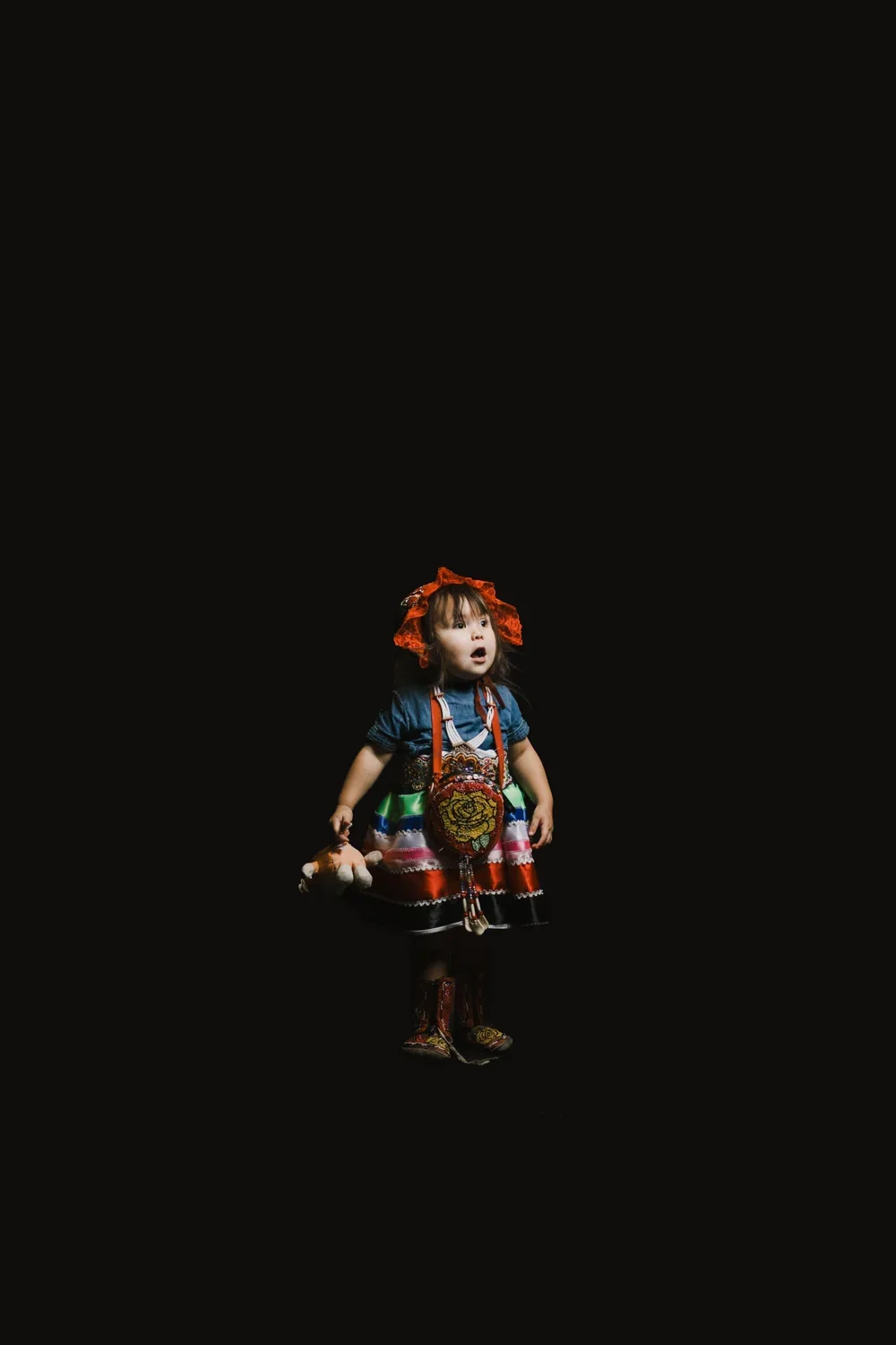 A young girl dressed in traditional colorful clothing, standing against a black background, looking surprised or amazed.