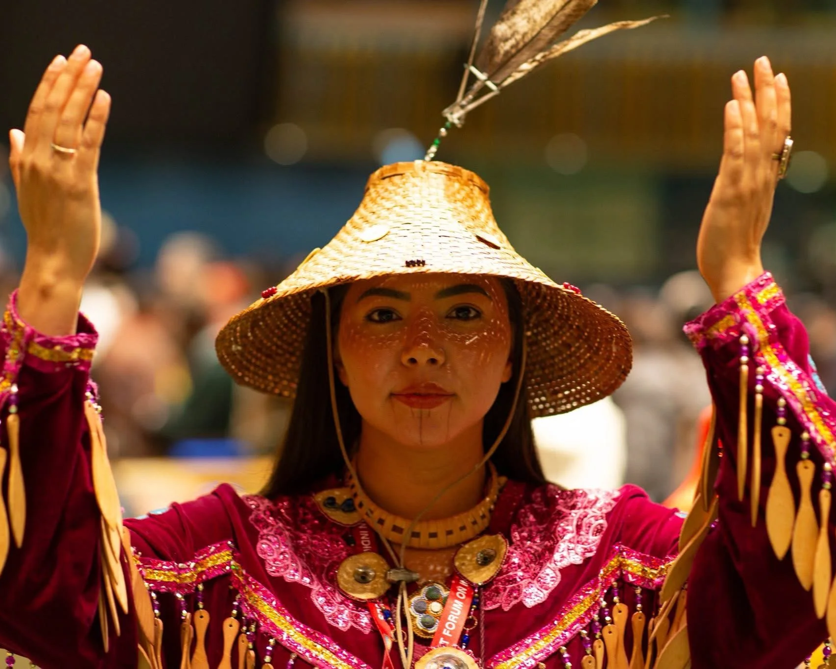 A woman wearing traditional indigenous attire, including a woven hat, with face paint and jewelry, holding a cultural artifact or performing a ritual at a festival or celebration.