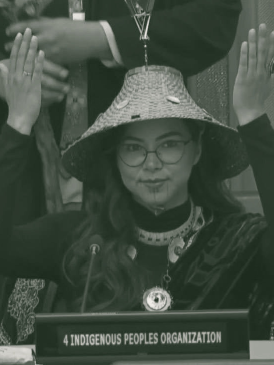 A woman wearing glasses and traditional Indigenous jewelry, sitting at a desk with a sign that reads '4 Indigenous Peoples Organization,' wearing a large woven hat, and holding her arms up with hands placed on the desk.