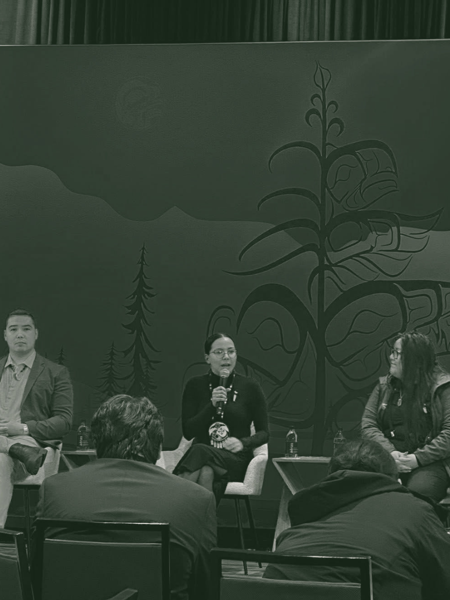 Panel discussion with three people on stage, one woman speaking into a microphone, in front of a mural of a plant and trees, audience seated in front.