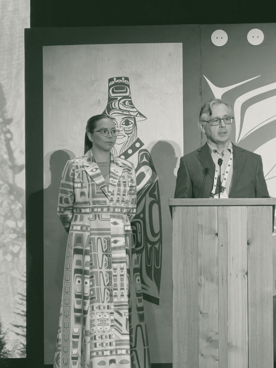 A woman and a man are standing at a podium during a presentation. The woman is wearing a patterned coat and glasses, and the man is wearing glasses and a suit. Behind them is artwork with indigenous designs and two circular symbols.