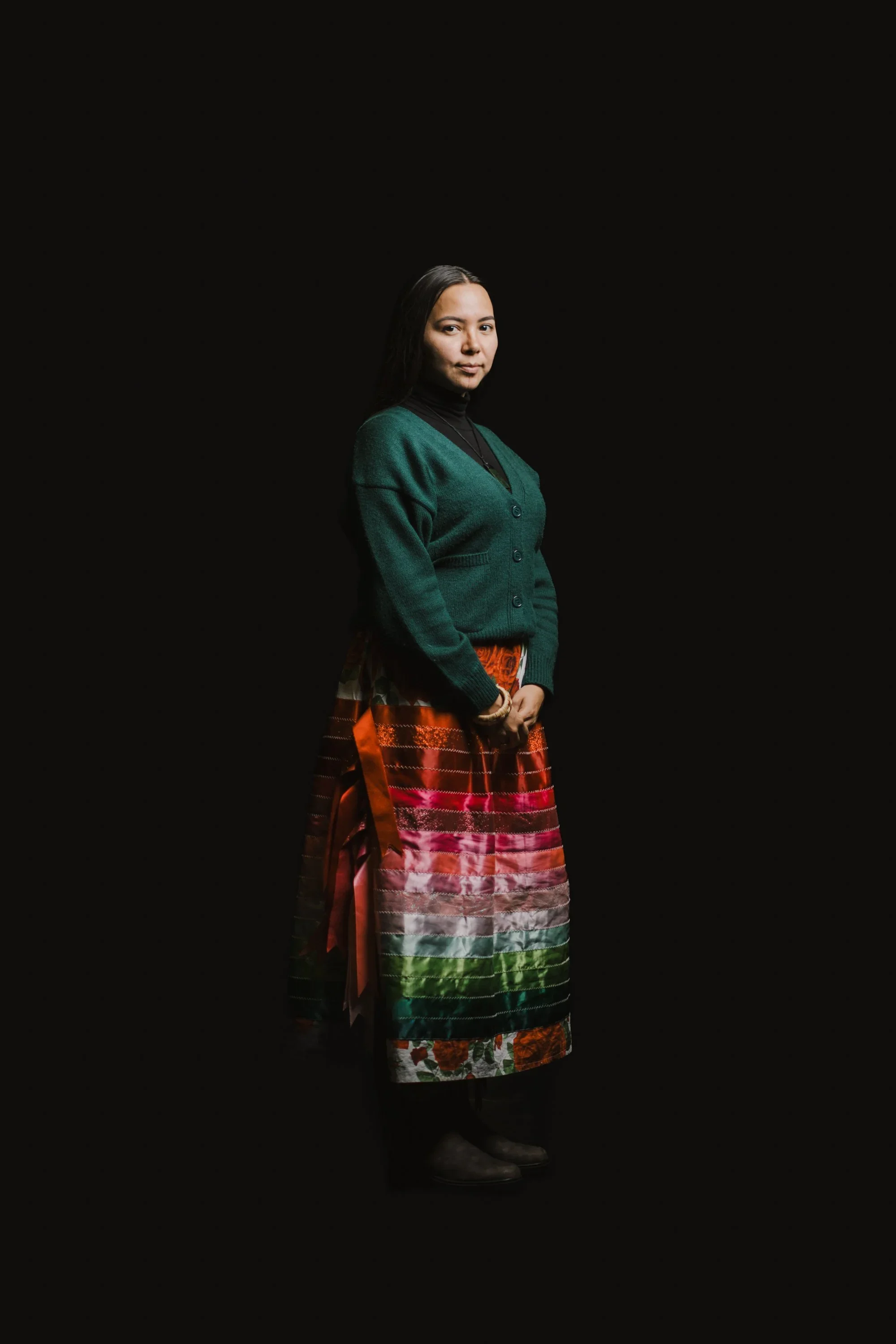 A woman standing against a black background wearing a green cardigan over a black turtleneck and a colorful, rainbow-striped skirt with floral patterns at the bottom. She has dark hair and is looking at the camera.