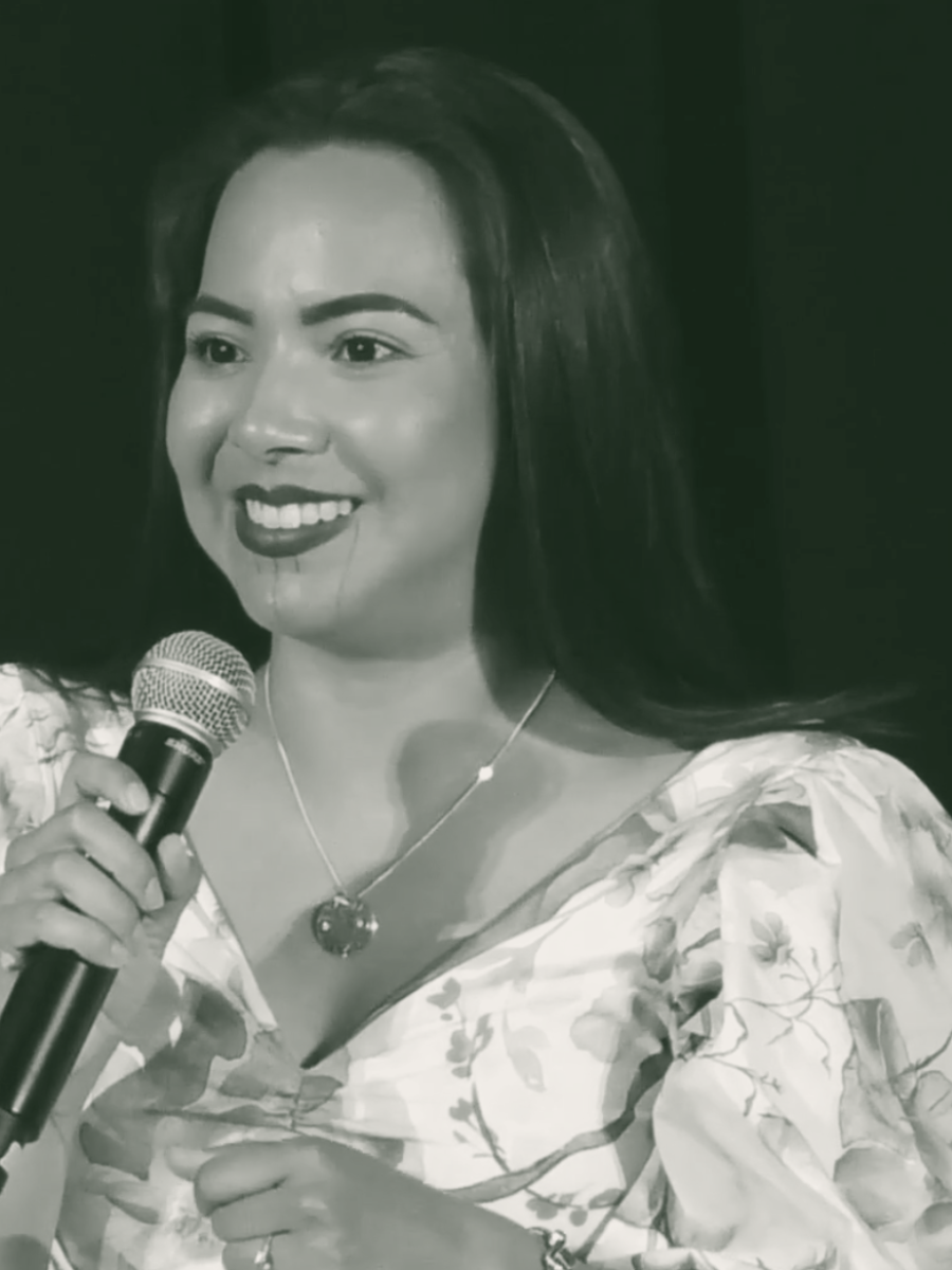 A woman with long dark hair smiling while holding a microphone and speaking, wearing a floral blouse and a necklace with a round pendant.