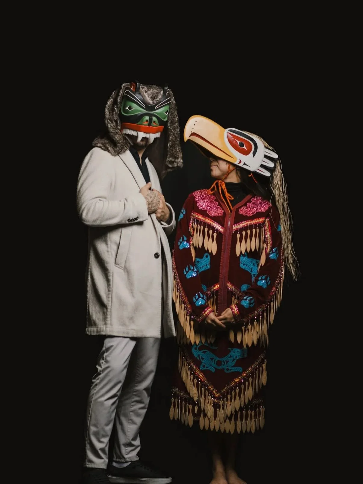 Two people wearing traditional masks and outfits stand against a black background, engaging with each other.