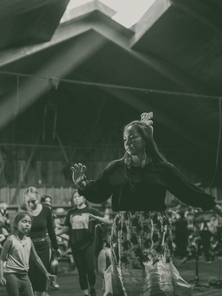 A woman with long hair and earrings appears to be leading a group of children in a dance or activity inside a large indoor space, like a gym or warehouse. The image is black and white.