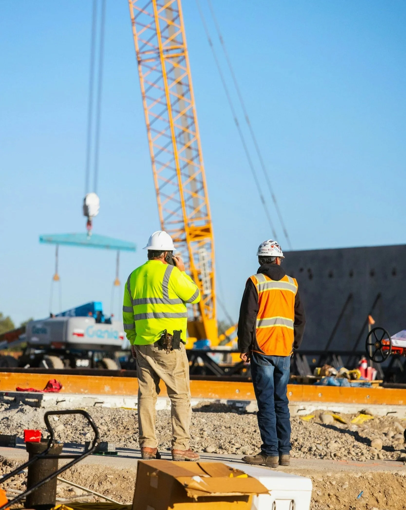 Industrial Construction site with two welders meeting to consult on a shutdown, one in a yellow safety jacket and the other in an orange safety vest. A tall yellow crane is in the background, and construction equipment is visible around them.