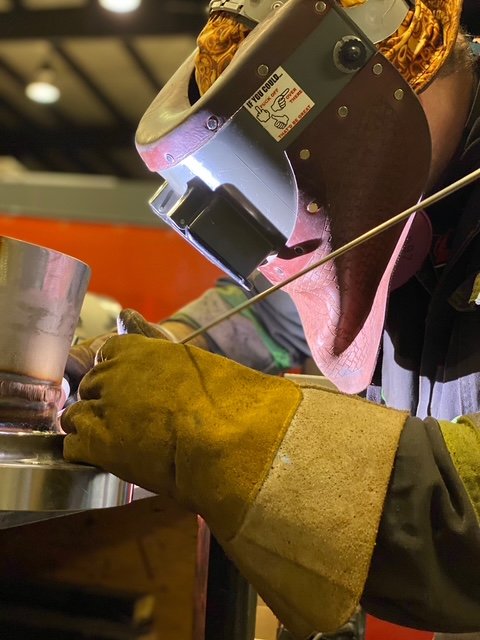 Person wearing protective welding gear, including a welding helmet and gloves, using a welding torch on a metal object.