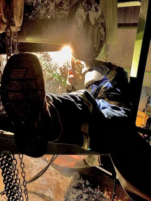 A person welding on site wearing a helmet and work gloves. Theynare working on a large piece of equipment in a workshop.