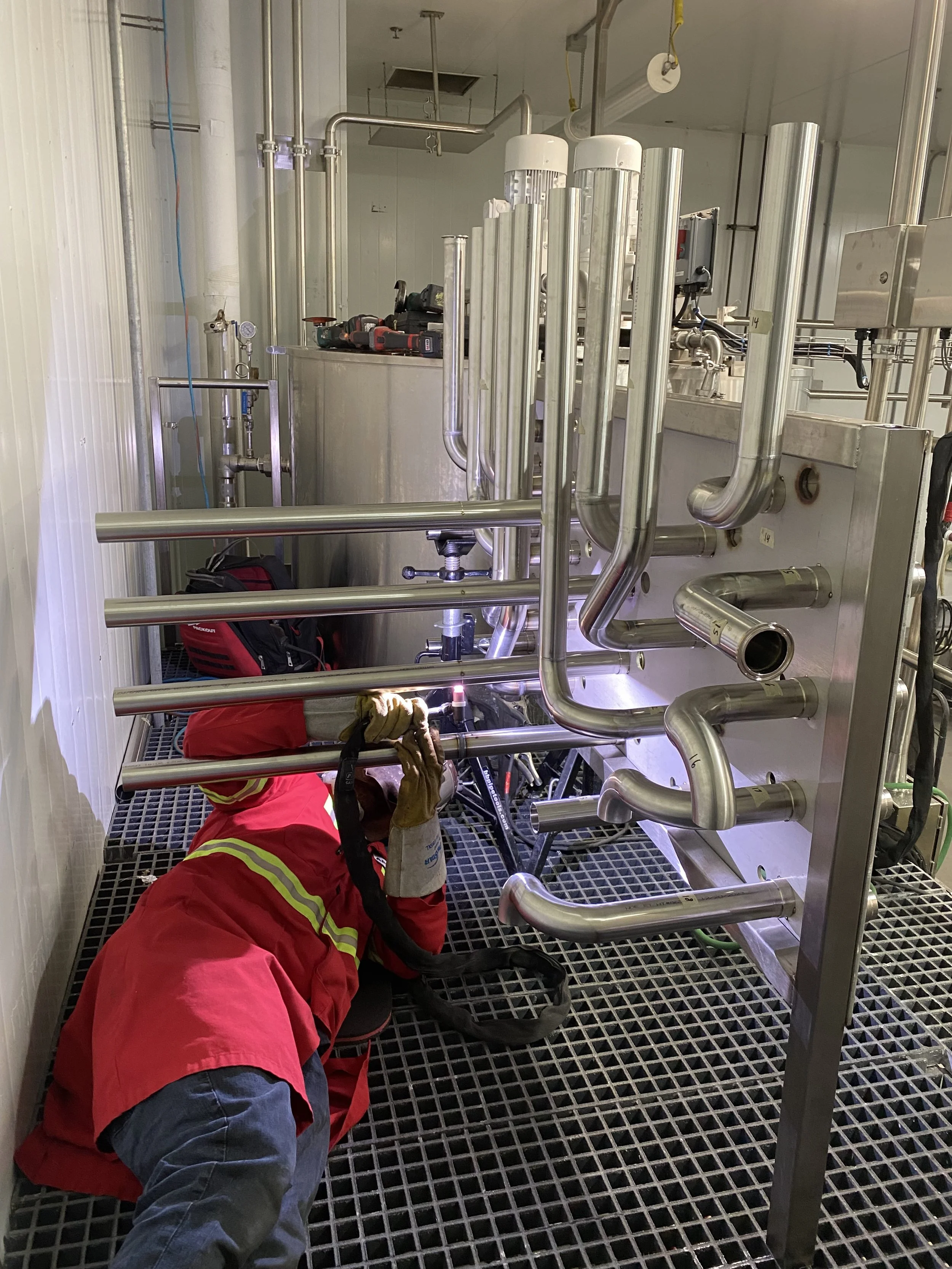 A worker in a red safety jacket and gloves is lying on the floor, welding and working on stainless steel pipes in an industrial setting. The workspace has metal grating flooring and various pipes and equipment.
