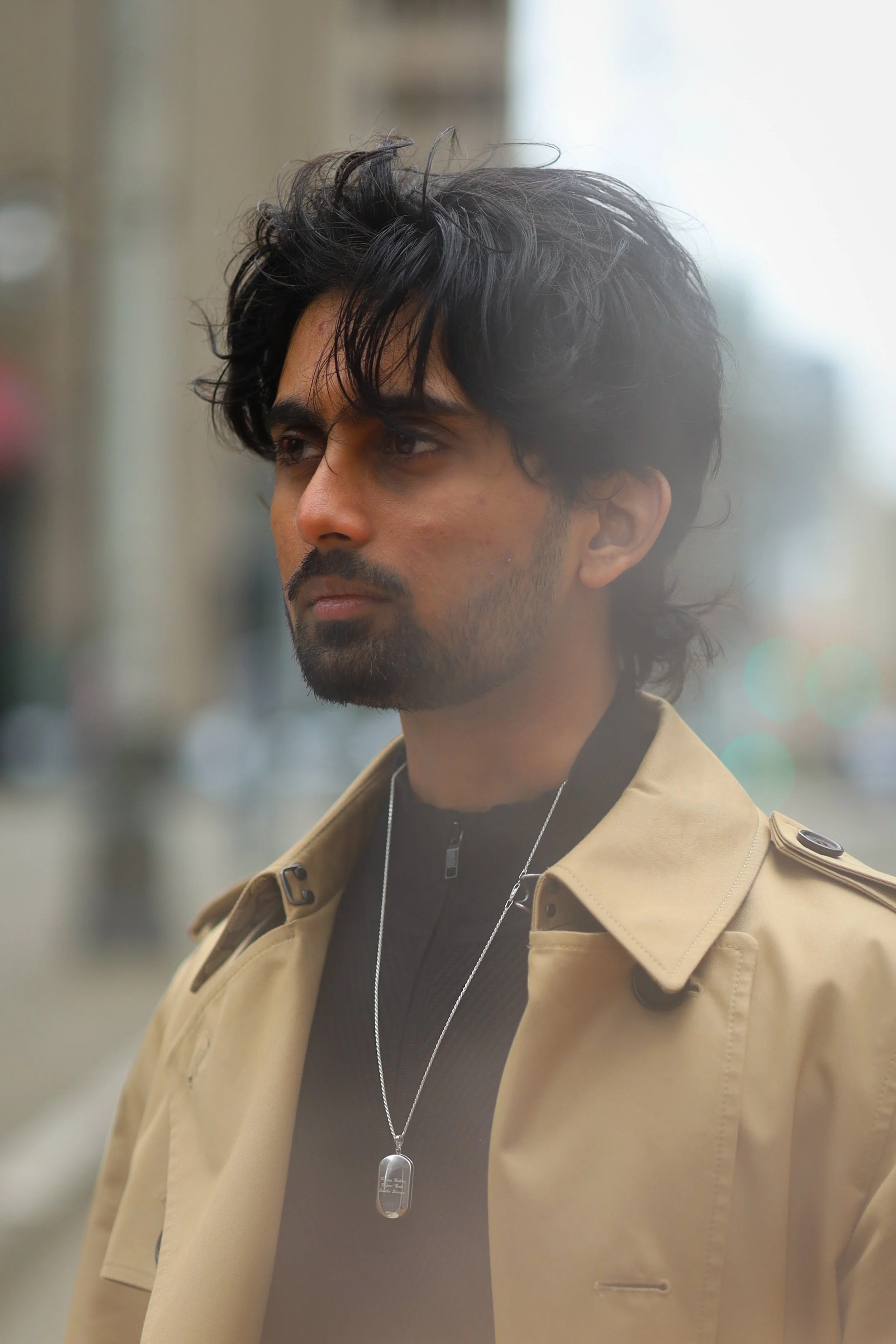 A young man with dark, wavy hair and a beard, wearing a beige coat with epaulets on the shoulders, a black shirt, and a silver dog tag necklace, standing outdoors in an urban setting.