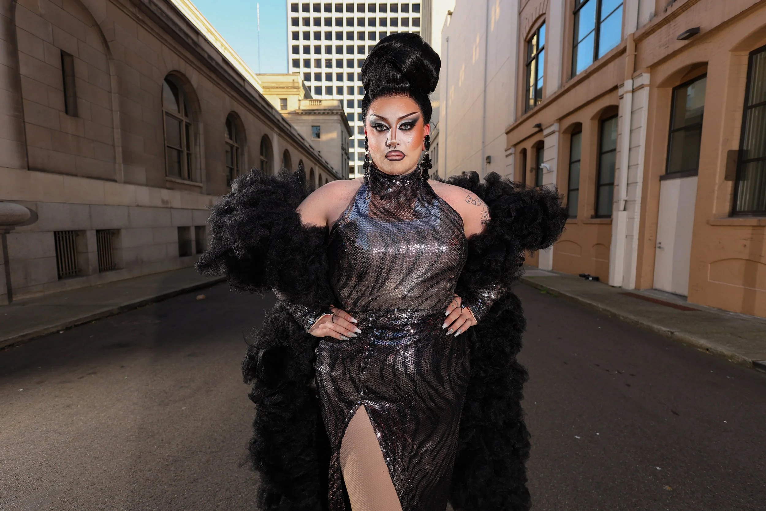 A drag queen dressed in a black sequin gown with a high slit, black fluffy sleeves, and large earrings, standing in an urban alleyway with tall buildings around.