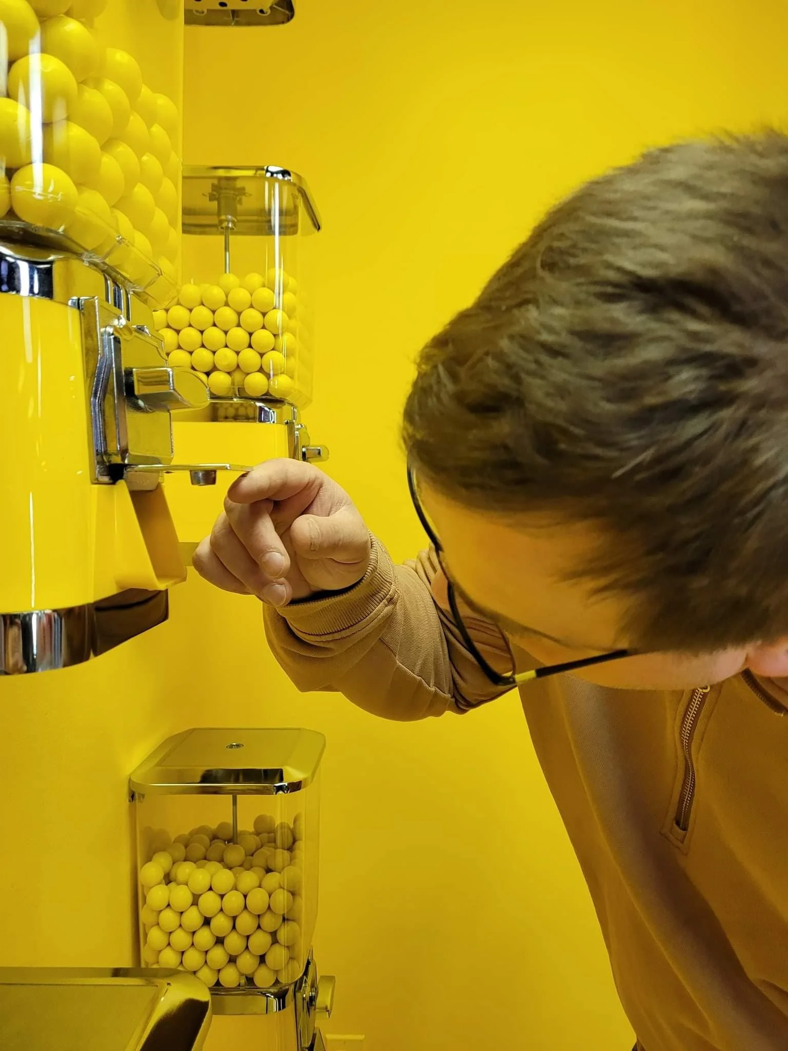A person with glasses and short hair is operating a yellow gumball machine filled with yellow gumballs, against a matching yellow wall.