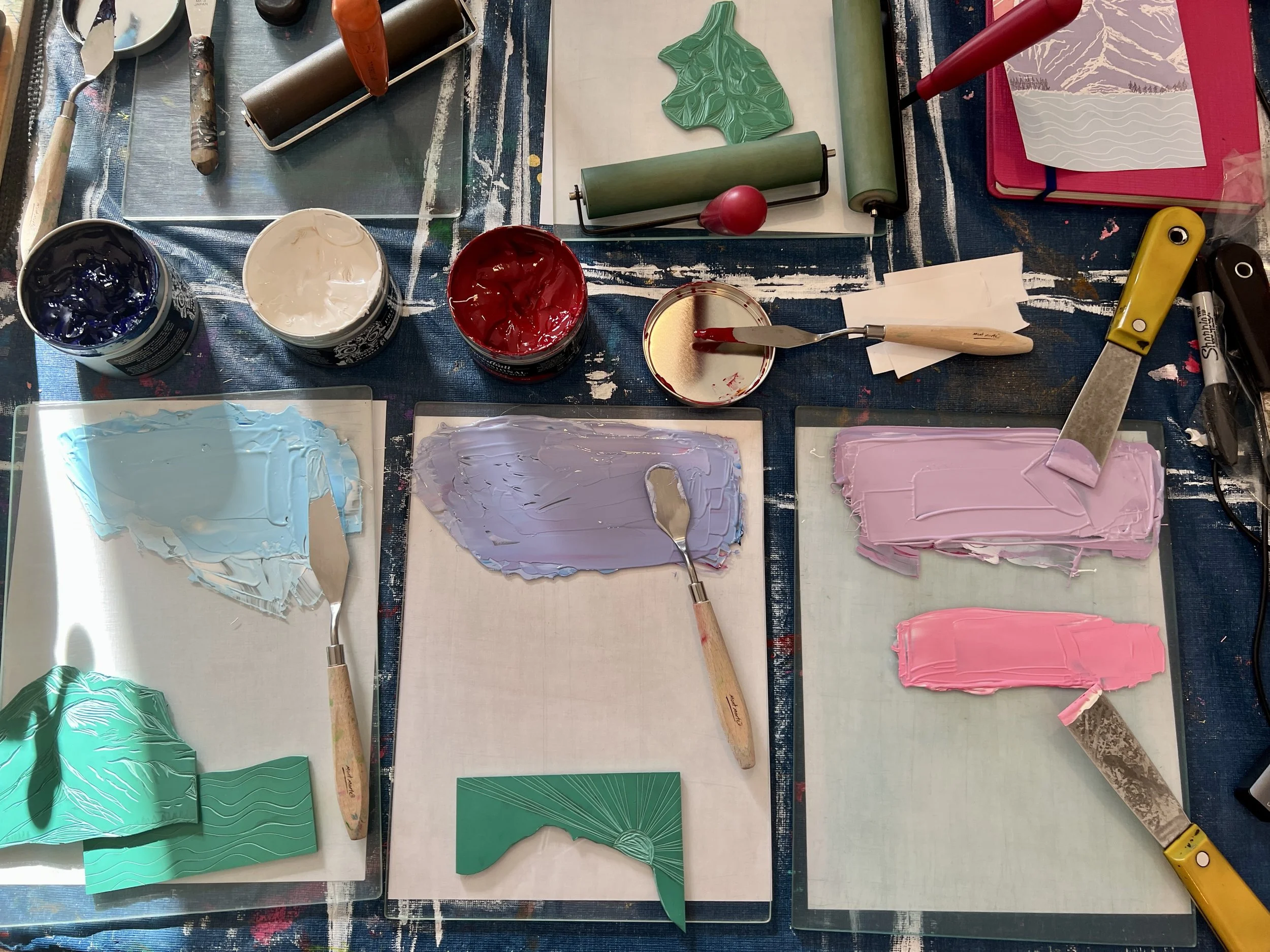 Studio workspace with printmaking supplies, including open tins of blue, white, and red ink, brayers, spatulas, and carved linocuts, ink in pink, purple, and blue are rolled out on glass slabs on top of a splattered table.