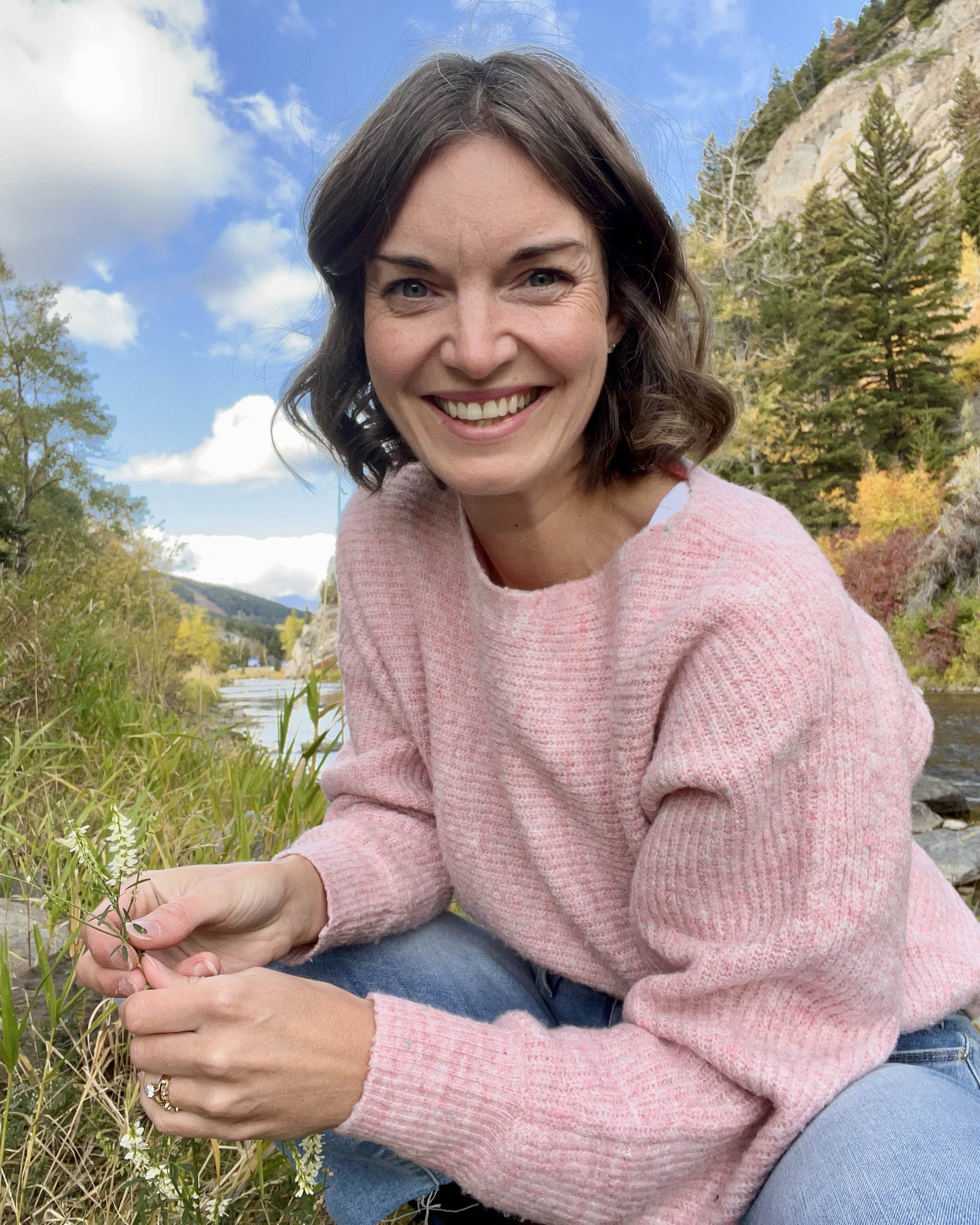 A smiling women with short brown hair is holding a plant by a river in the fall in the mountains