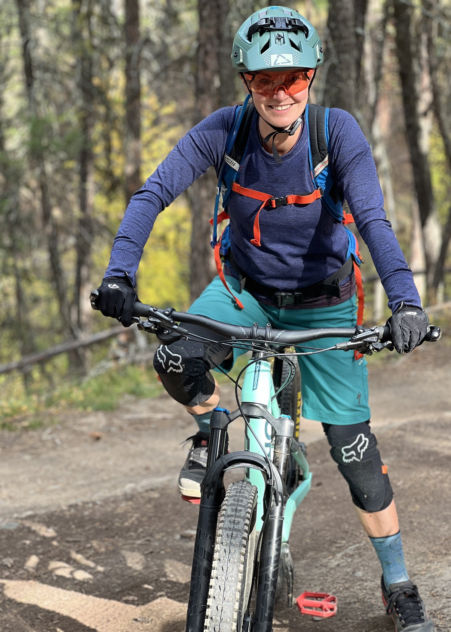 A woman mountain biking on a trail in a forest, wearing a helmet, sunglasses, covered arms, a backpack, and riding gear, smiling at the camera.