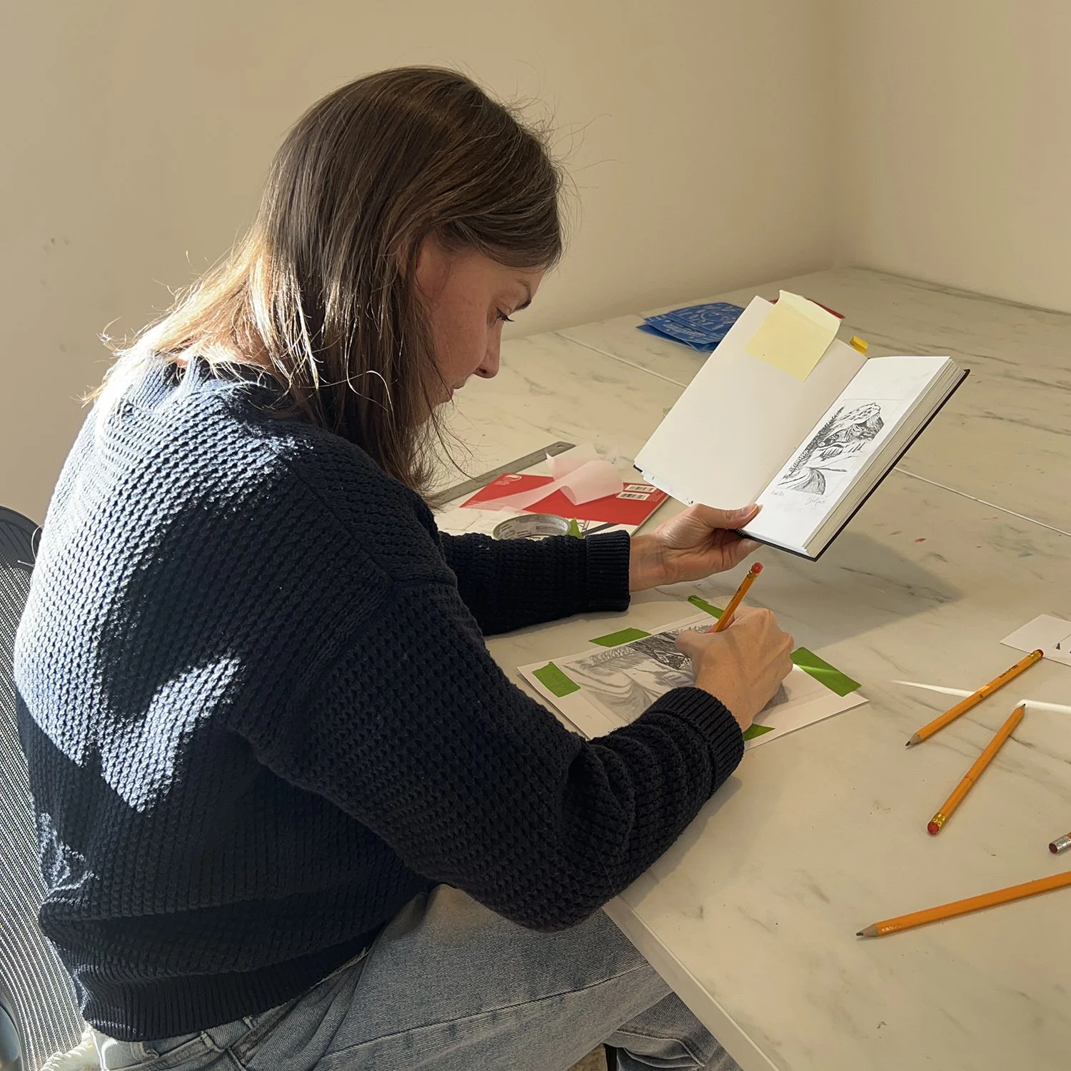 A woman with brown hair sitting at a white table, drawing black and white nature scenes on paper with pencils and looking at a sketchbook.