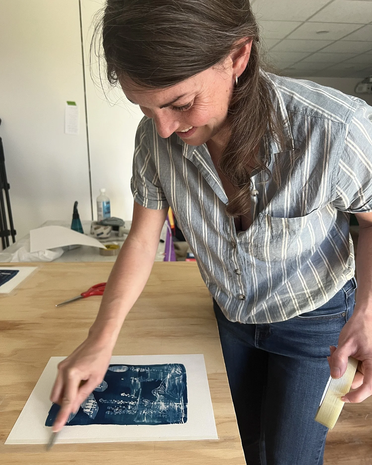 A woman leaning over a table, rubbing down a blue and white artwork, with scissors, a roll of tape, and documents on the table in a room with a plain wall and a white cabinet.