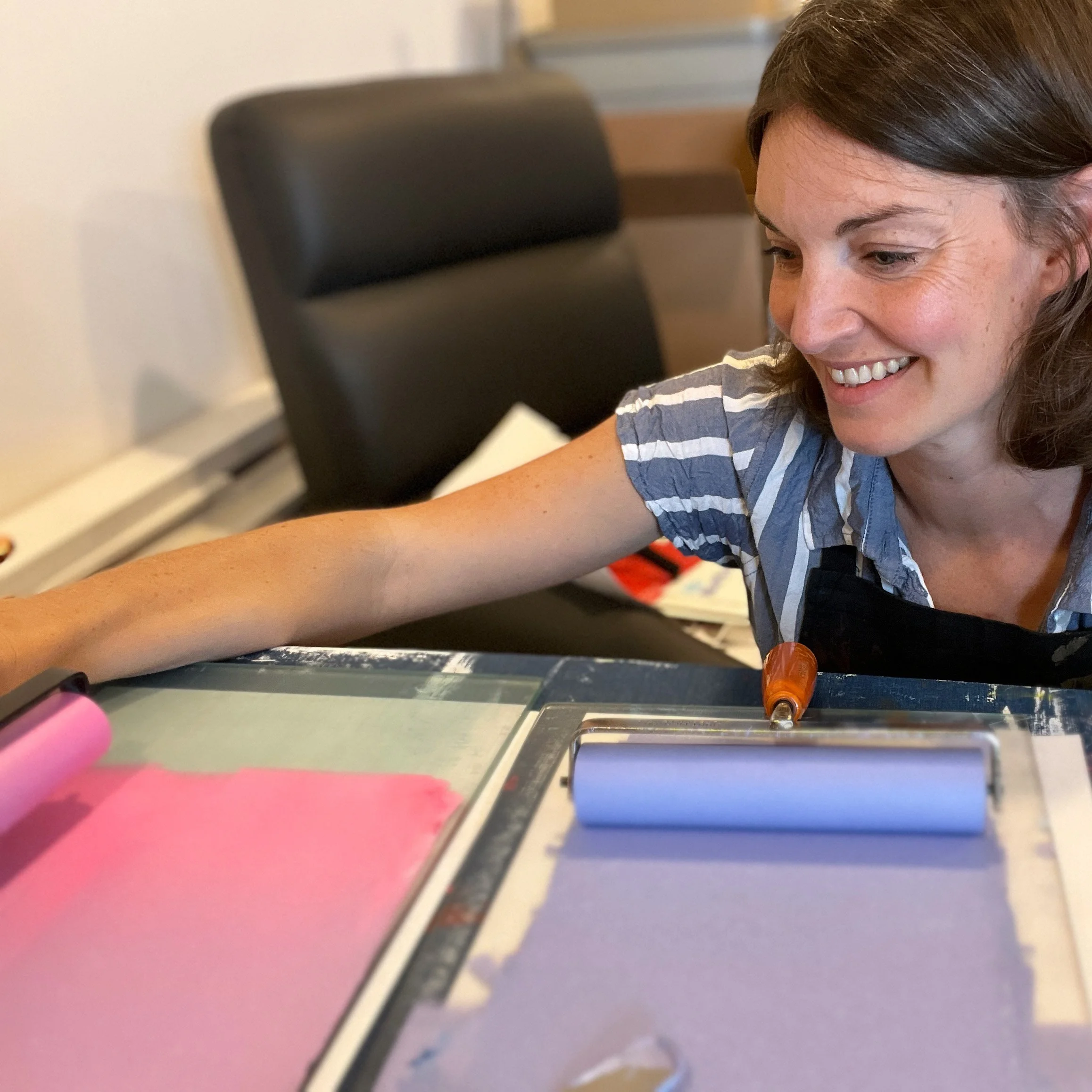 Close-up of a women's face, smiling, with dark hair, wearing a shirt with blue and white stripes, sitting at a desk with pink and purple printmaking ink visible in the foreground.