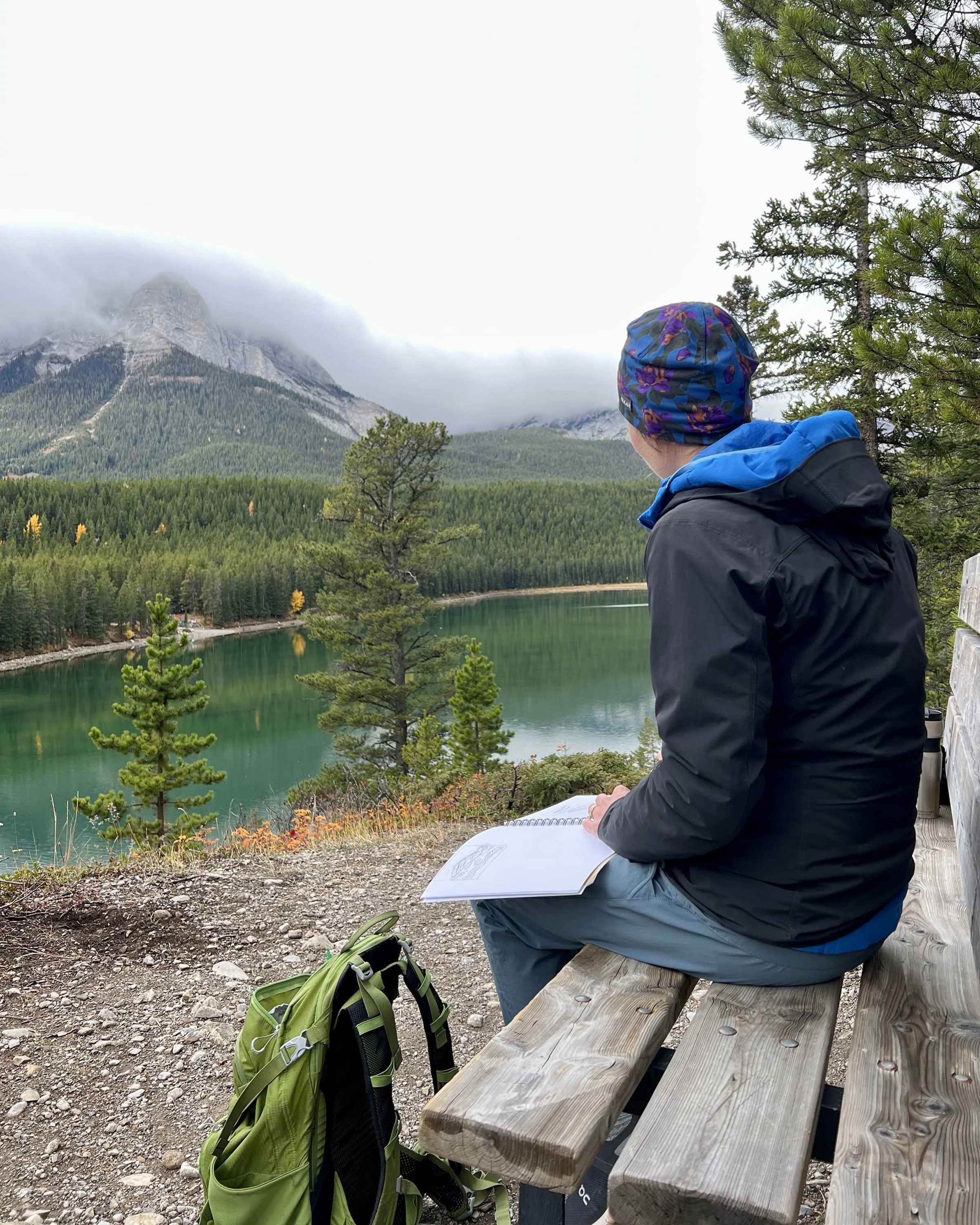 Women sitting on a wooden bench sketching and overlooking a mountain lake with forested hills and a mountain peak partially covered in clouds, surrounded by trees and foliage.