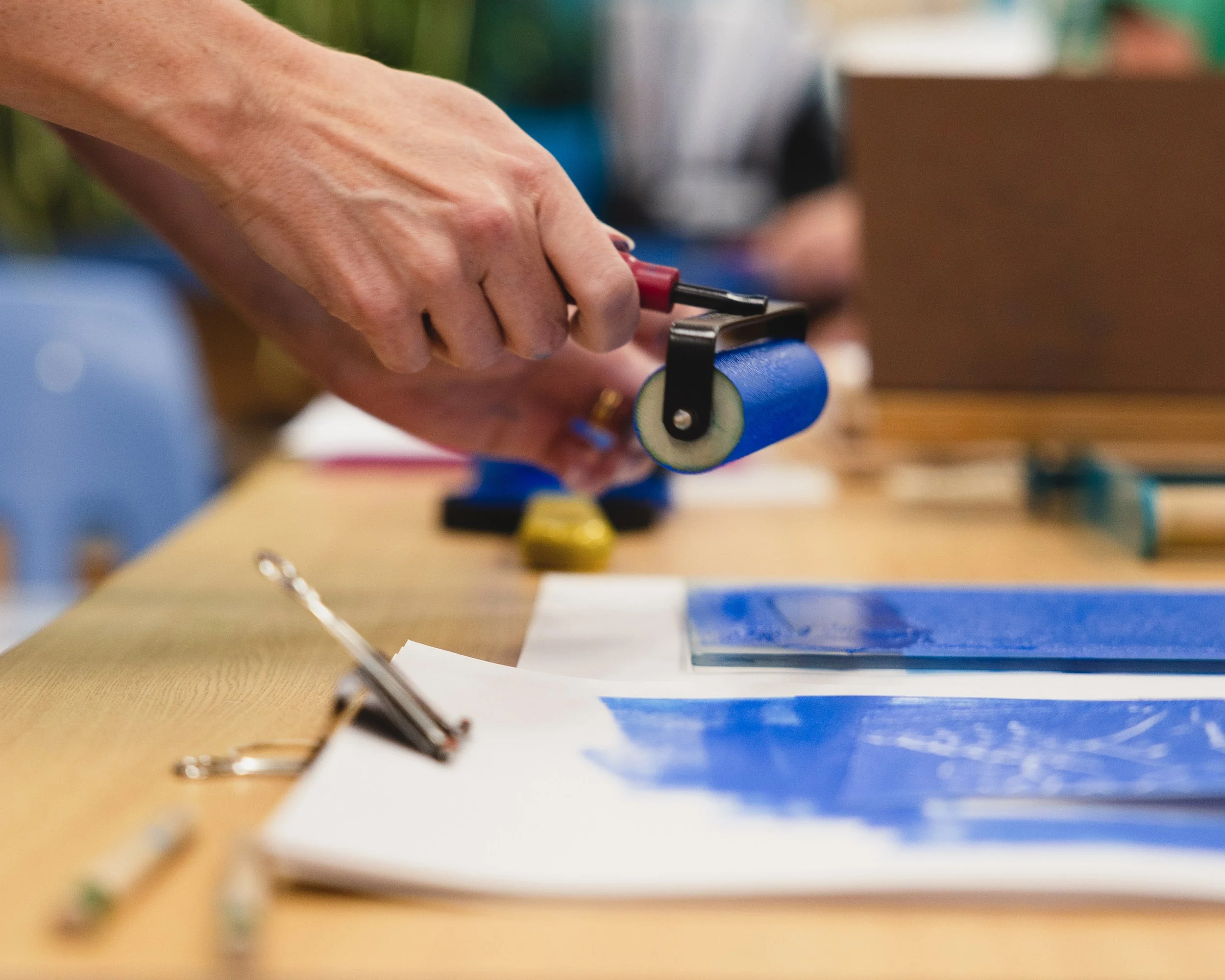 Women holding a brayer coated in blue ink above a glass slab and carved image that both also have blue ink. There is a printing press in the background.
