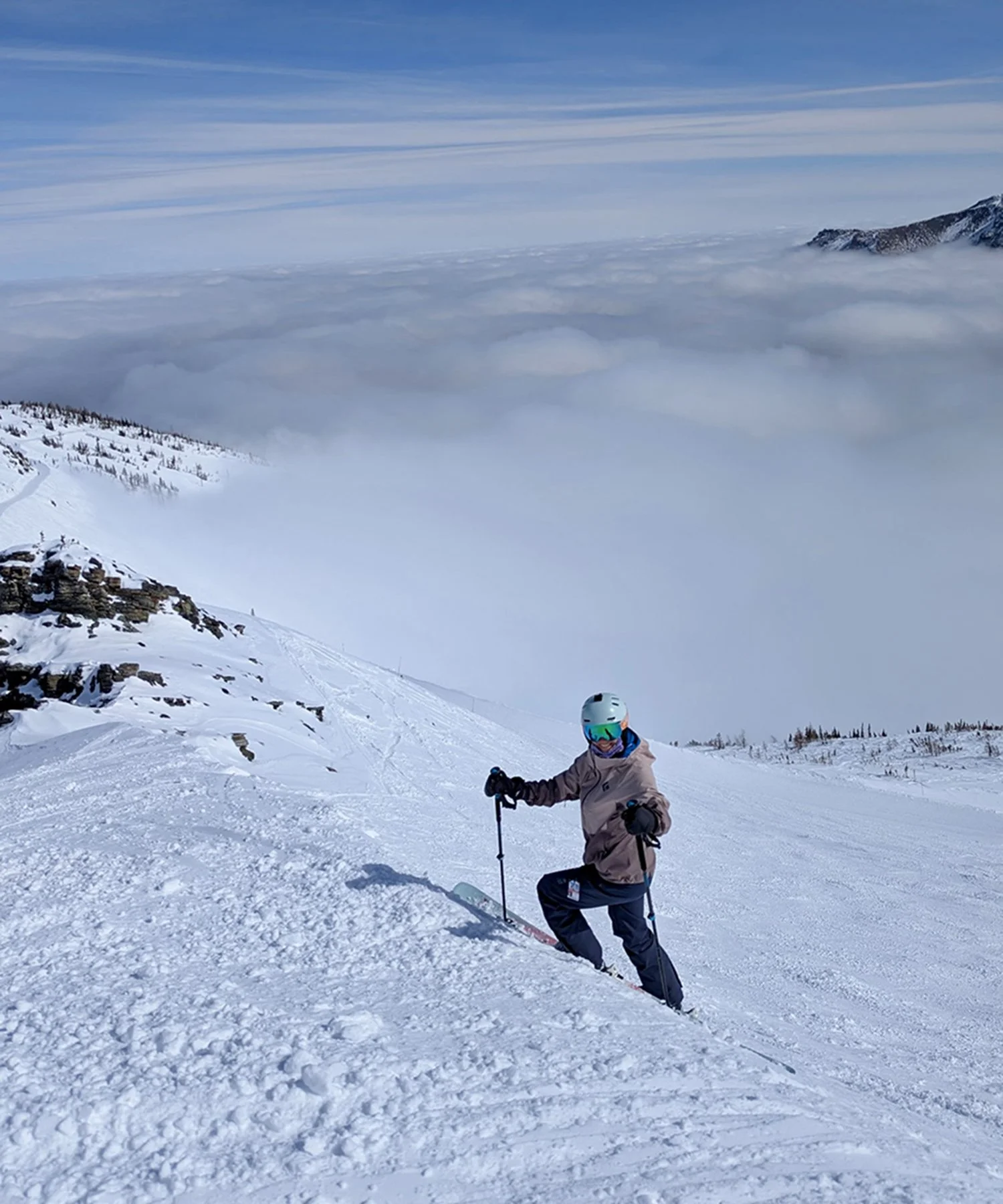 A women wearing a ski helmet and goggles stands on a snowy mountain slope, holding ski poles. The mountain is covered in snow, with a rocky outcrop on the left, and a blanket of clouds covering the landscape below. The sky is clear with some streaks 