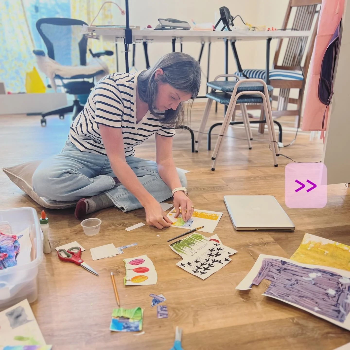 A woman sitting cross-legged on the floor working on art projects with various papers, scissors, and art supplies around her, in a bright room with a table and chairs in the background.