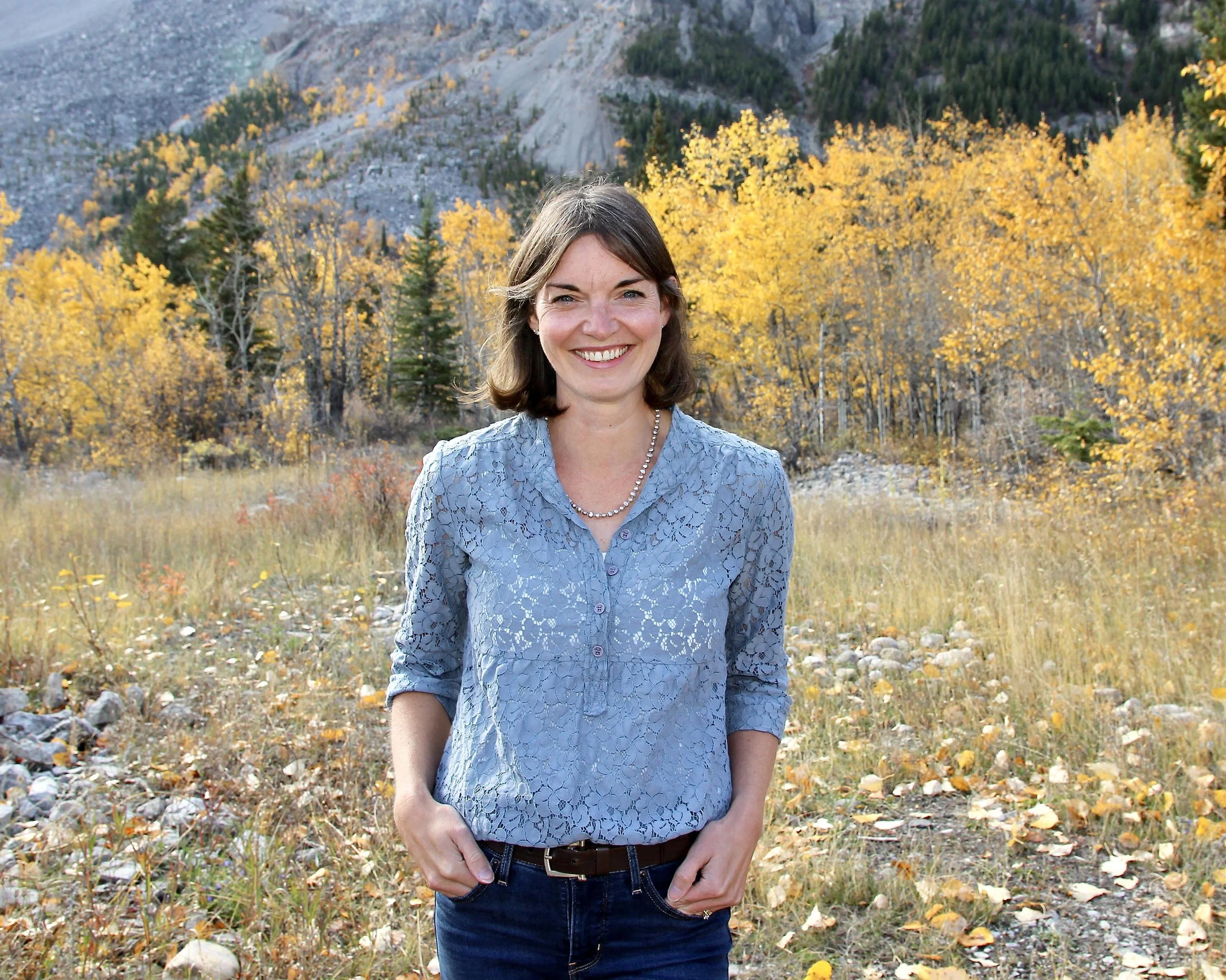 A woman smiling outdoors with a backdrop of yellow and green autumn trees and a mountainside.