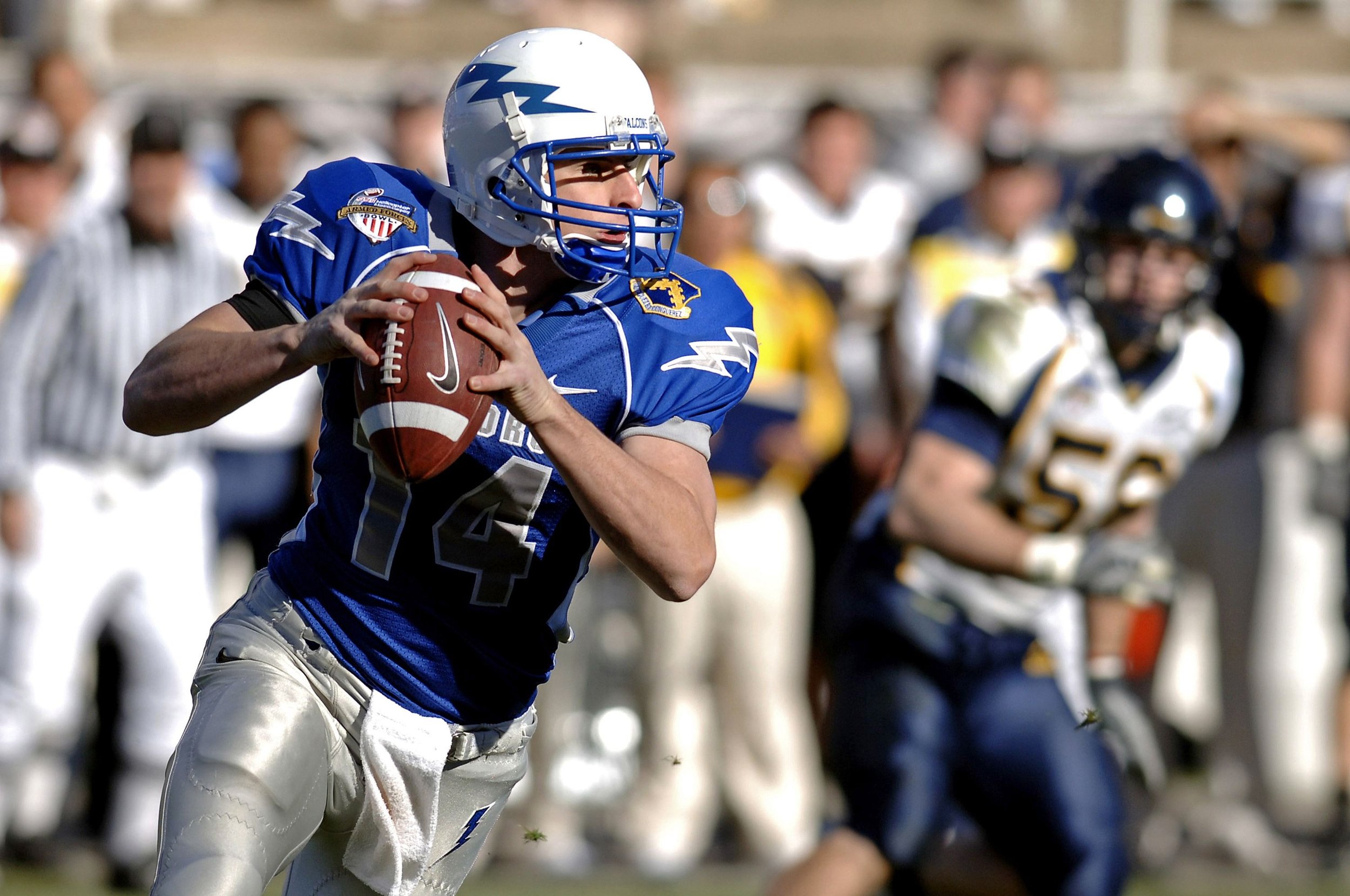 A football player in a blue jersey and white helmet holding a football, running during a game with players and officials in the background.