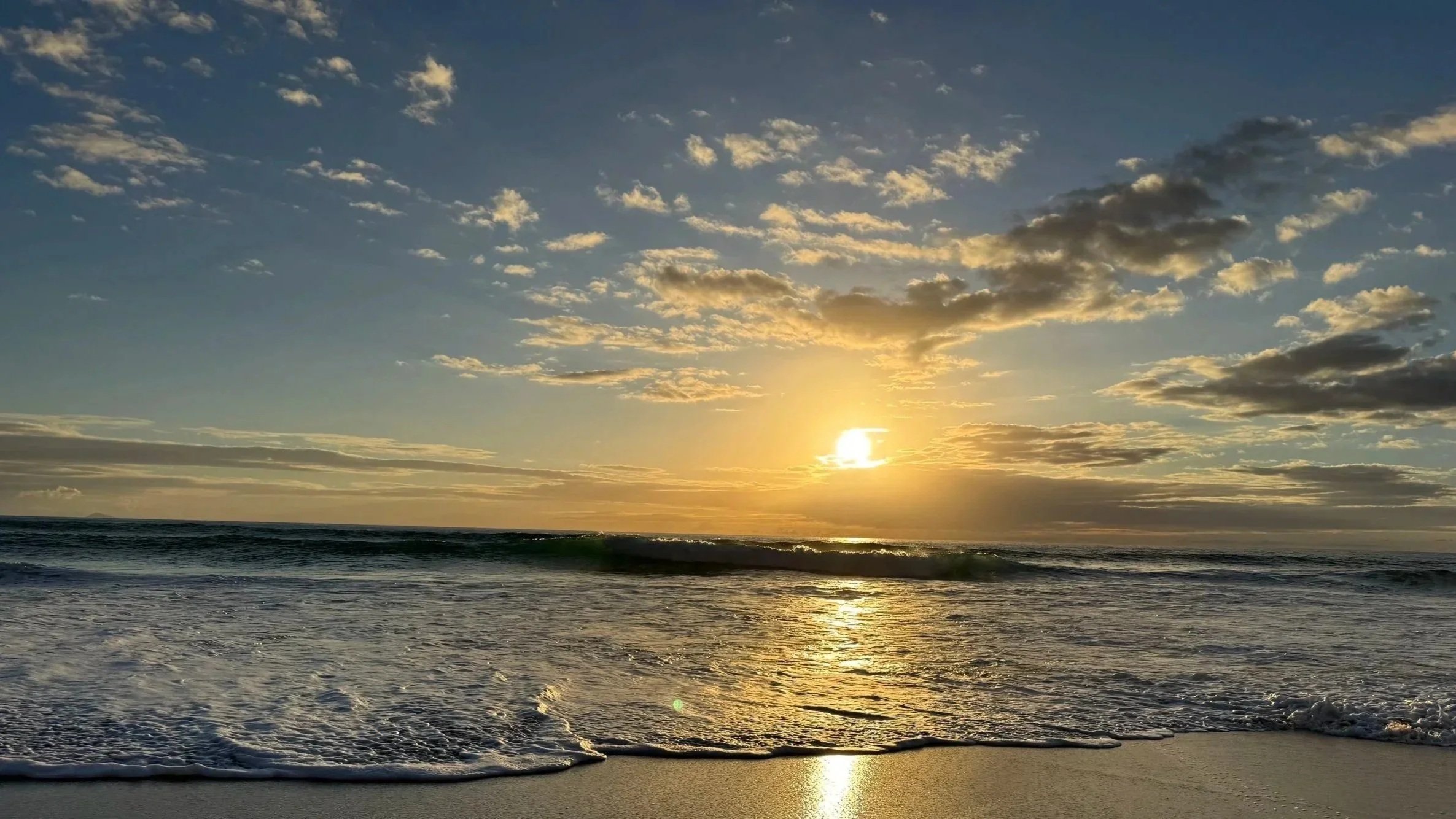 Sun setting over the ocean with clouds in the sky and waves crashing on the sandy beach.