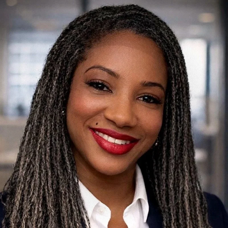 A woman with long braided hair, wearing lipstick, a white collared shirt, and a dark blazer, smiling in an indoor setting.