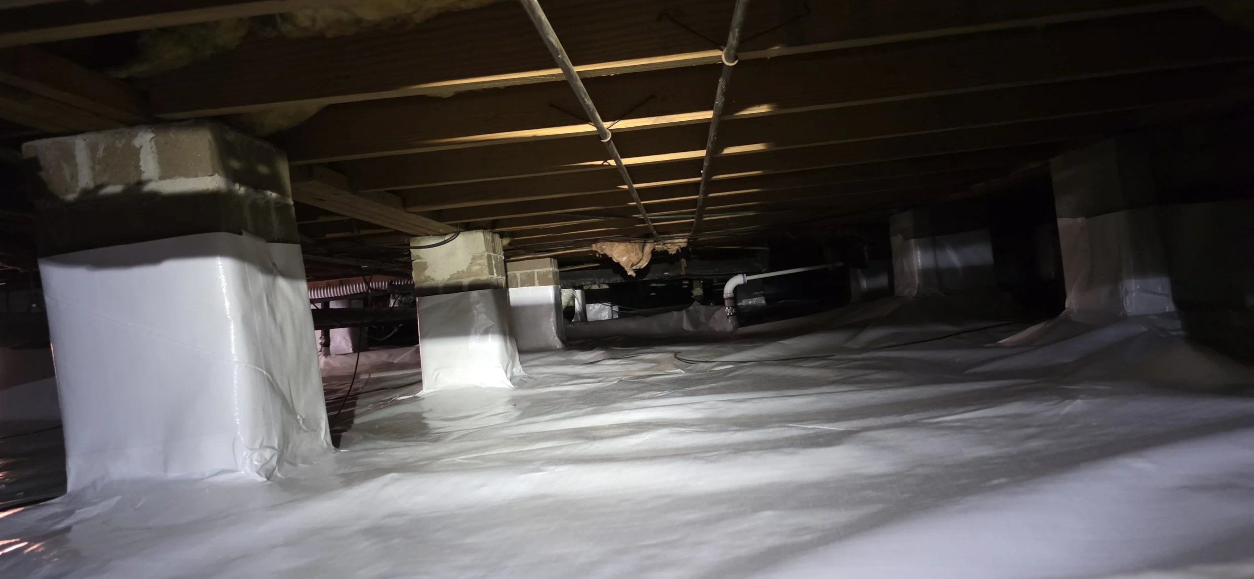 crawlspace with exposed ceiling joists, concrete support columns wrapped in white insulation, and plastic vapor barrier on the floor.