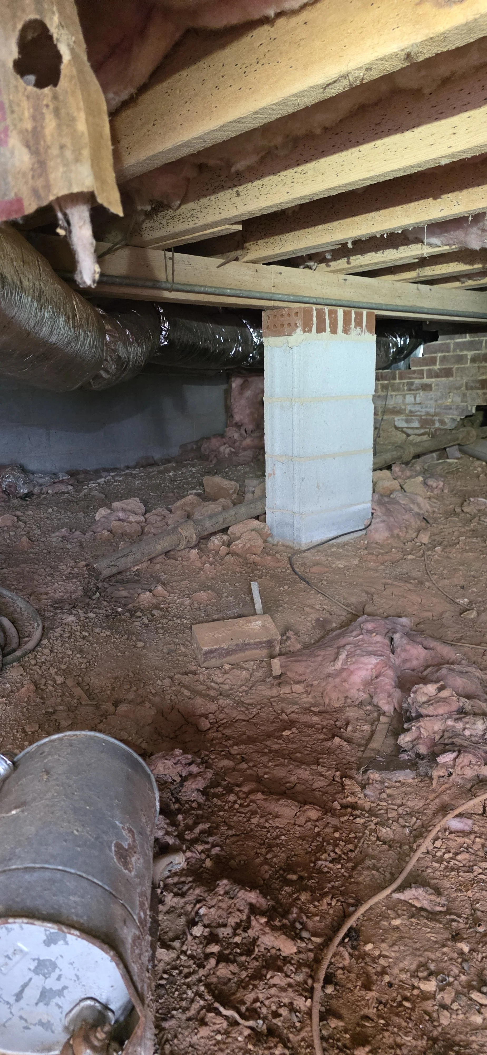 View of a construction site crawlspace with exposed soil, insulation, pipes, and a brick and concrete pillar supporting the floor above, indicating ongoing building or renovation work.