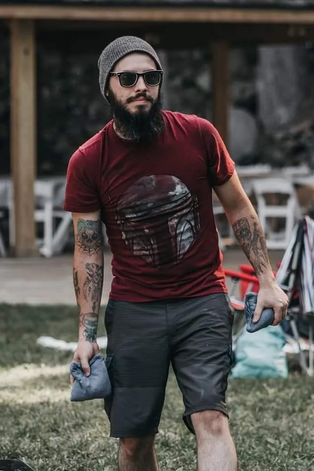 A man with a beard wearing sunglasses, a gray beanie, a red graphic t-shirt, and black shorts, holding gray cloths in both hands, standing outdoors on grass.