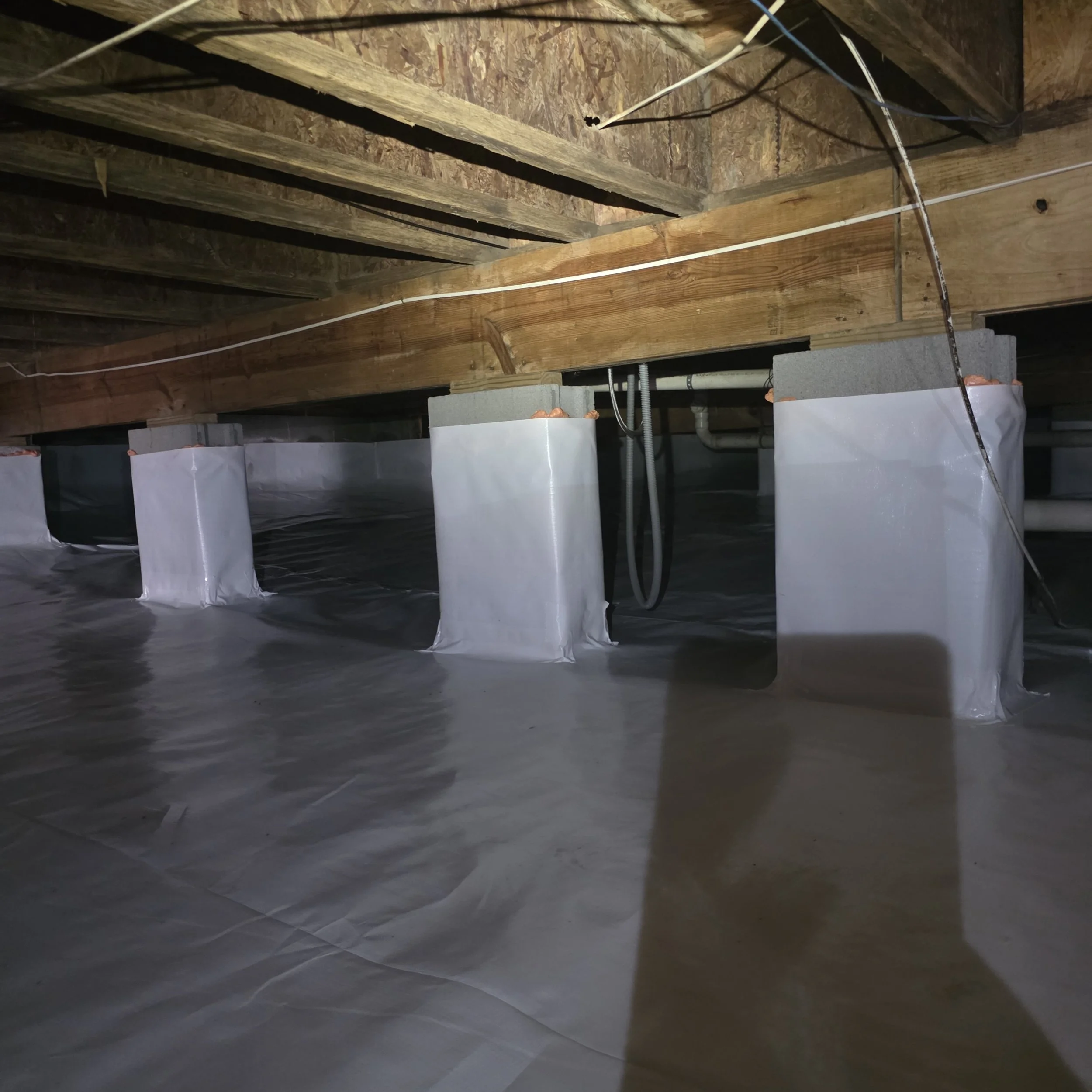 View of a crawlspace ceiling with exposed wooden beams, electrical wiring, and pipes, supported by concrete blocks and covered with a black vapor barrier on the floor.
