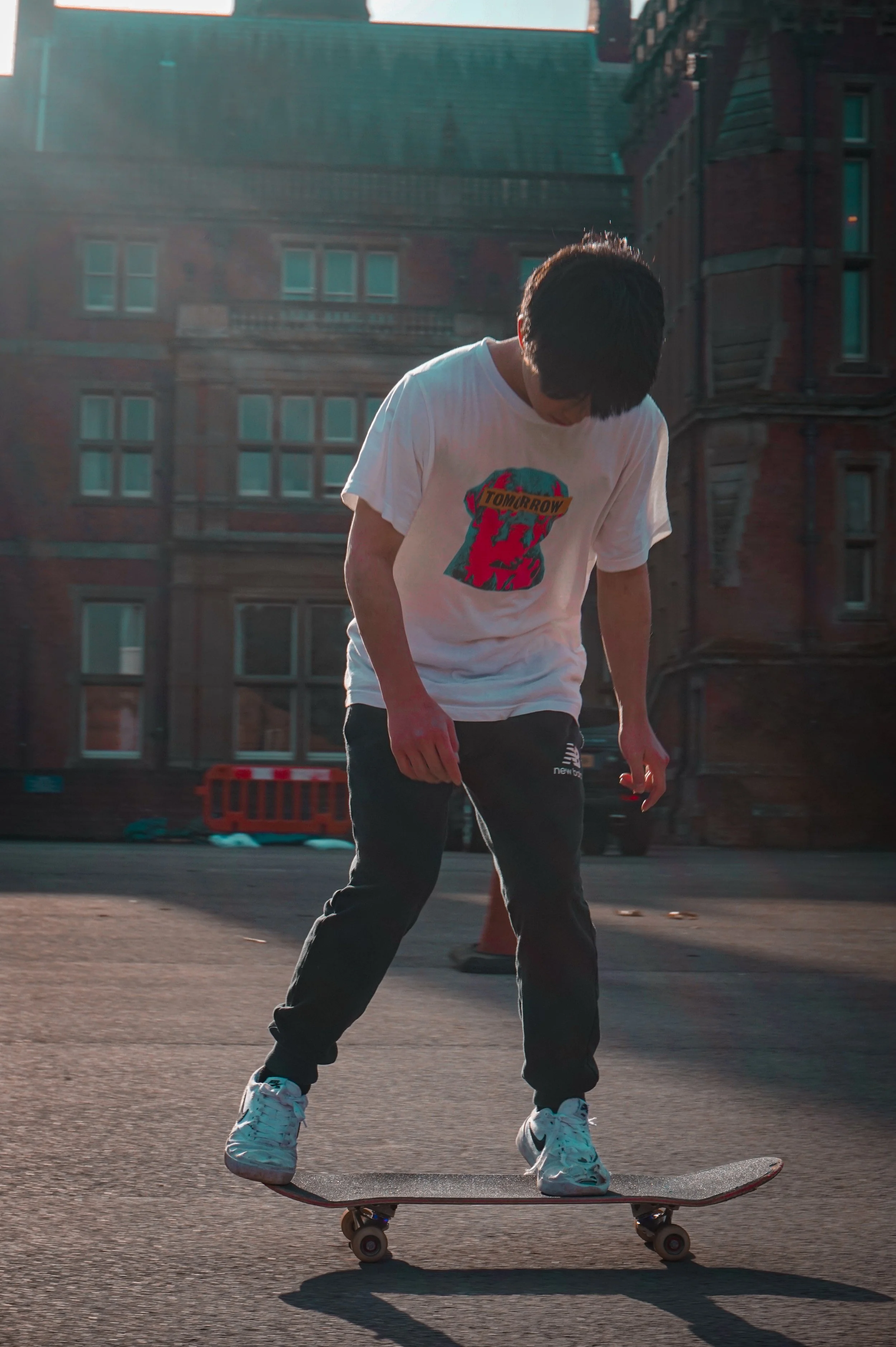 A young person skateboarding outdoors on a sunny day, wearing a white T-shirt with colorful graphics and text, dark pants, and white sneakers, with a brick building in the background.