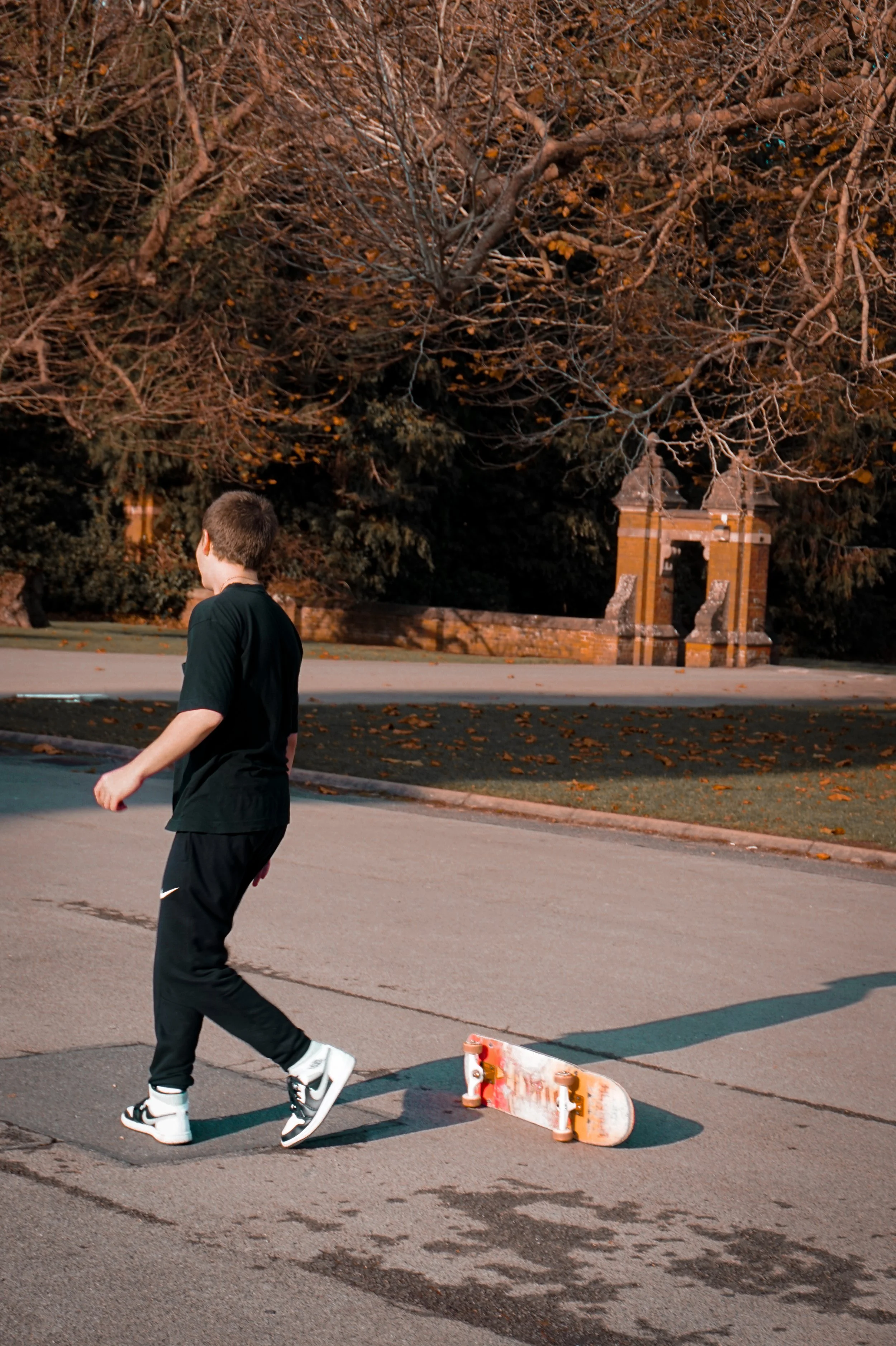 A person in black athletic clothing and sneakers walking past a skateboard on a paved area, with a background of large trees with autumn leaves and a brick structure archway.