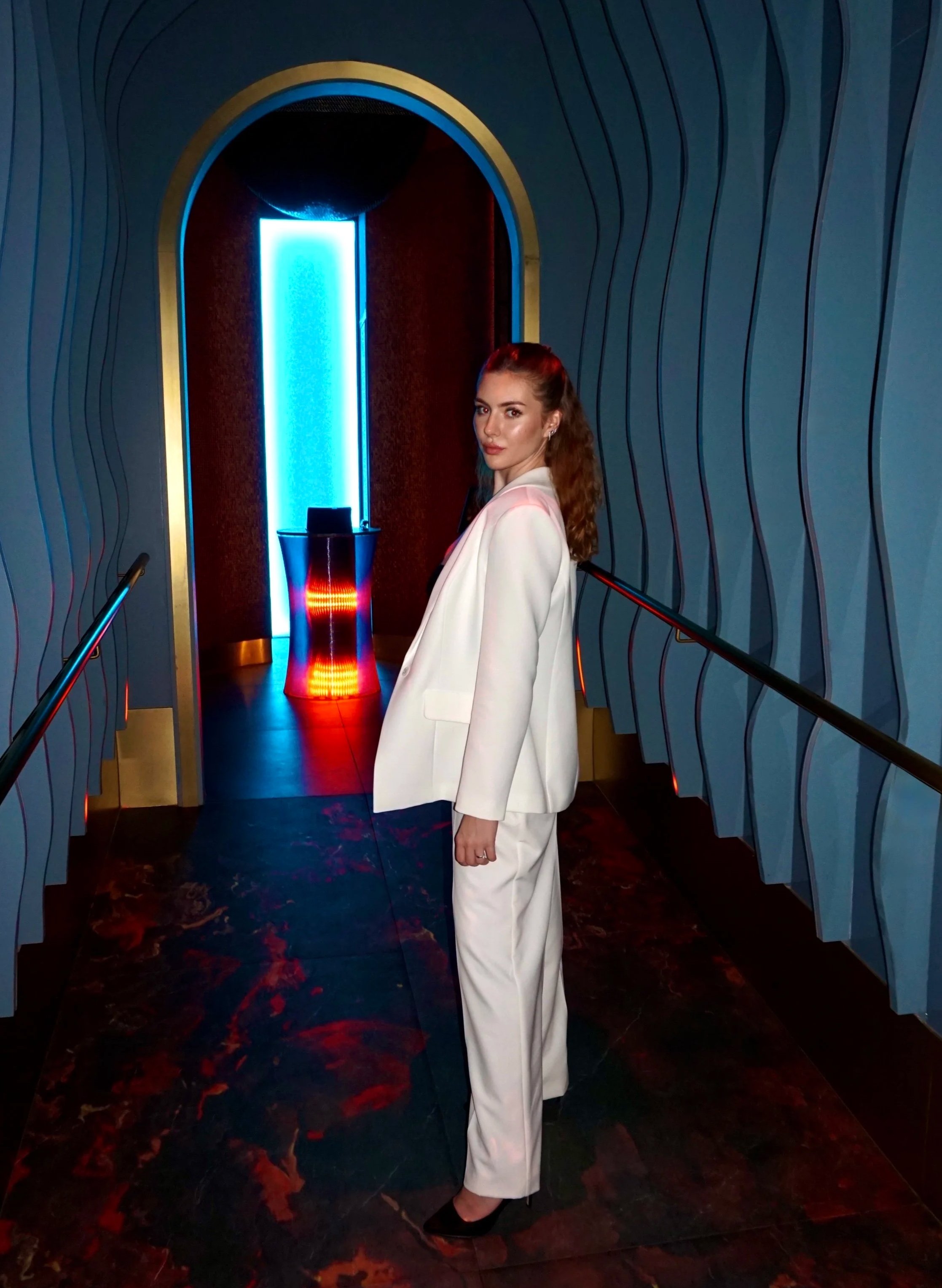 A woman in a white suit standing in a dimly lit corridor with blue and red neon lights, in front of a blue-lit door or screen.