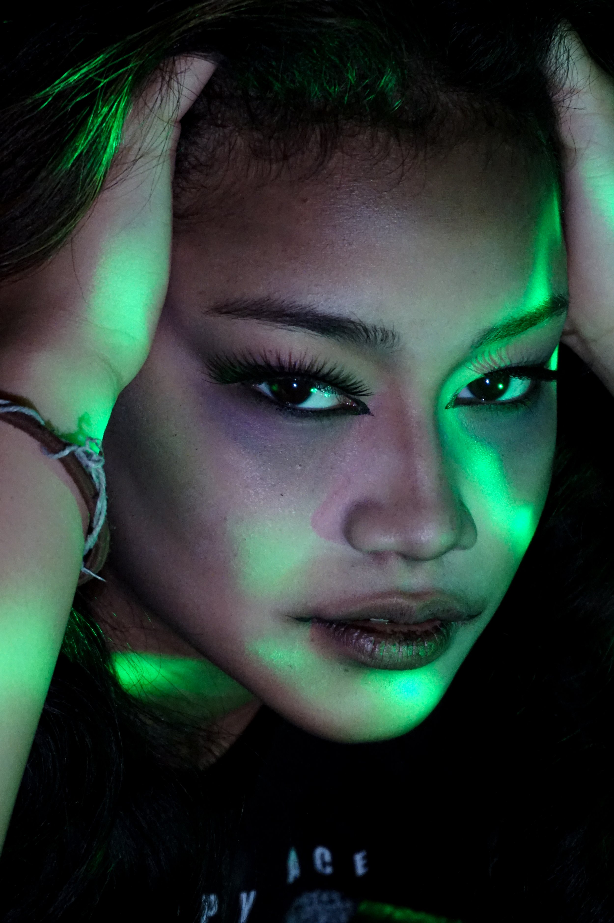 A close-up portrait of a person with long dark hair, distinctive eye makeup, and glowing green light effects on their face and hair, with a hand resting on their forehead.