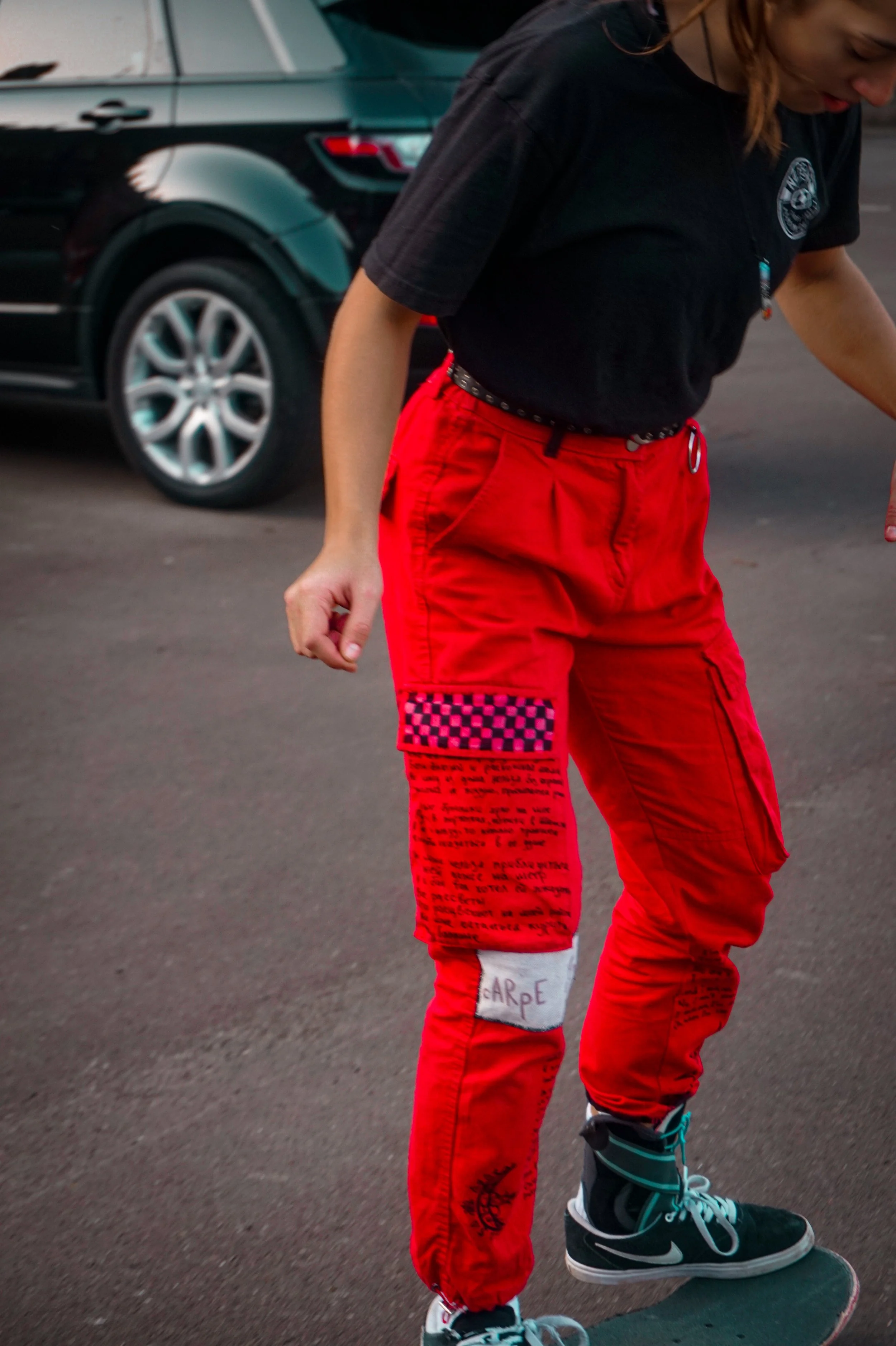 A person wearing red pants with text and graphic designs, a black shirt with a patch, and skating shoes riding a skateboard on a paved surface.