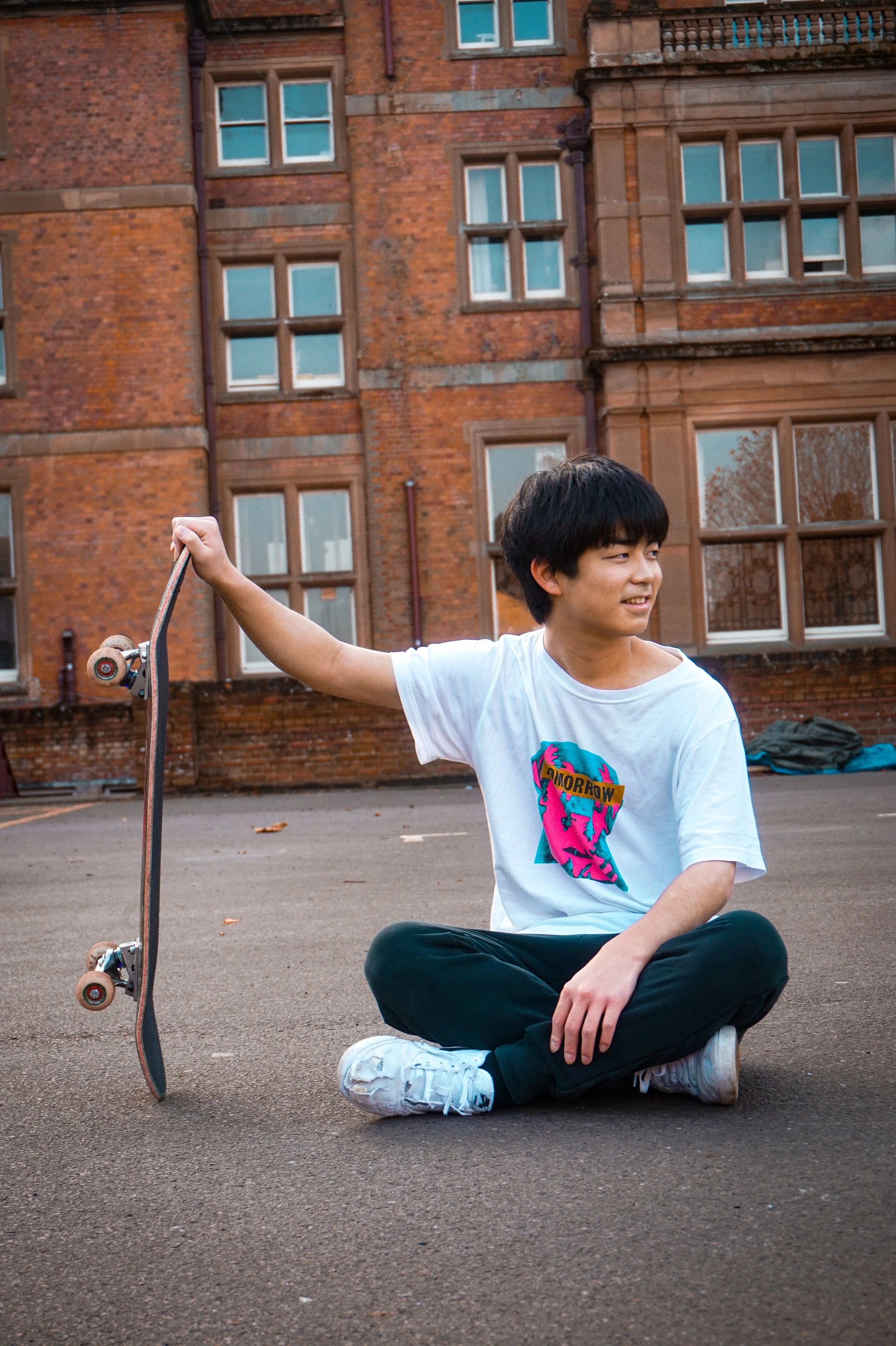 A young person with black hair, wearing a white t-shirt with a colorful graphic and the word 'Tomorrow,' sitting cross-legged on the ground outdoors, holding a skateboard in their right hand, with a brick building in the background.