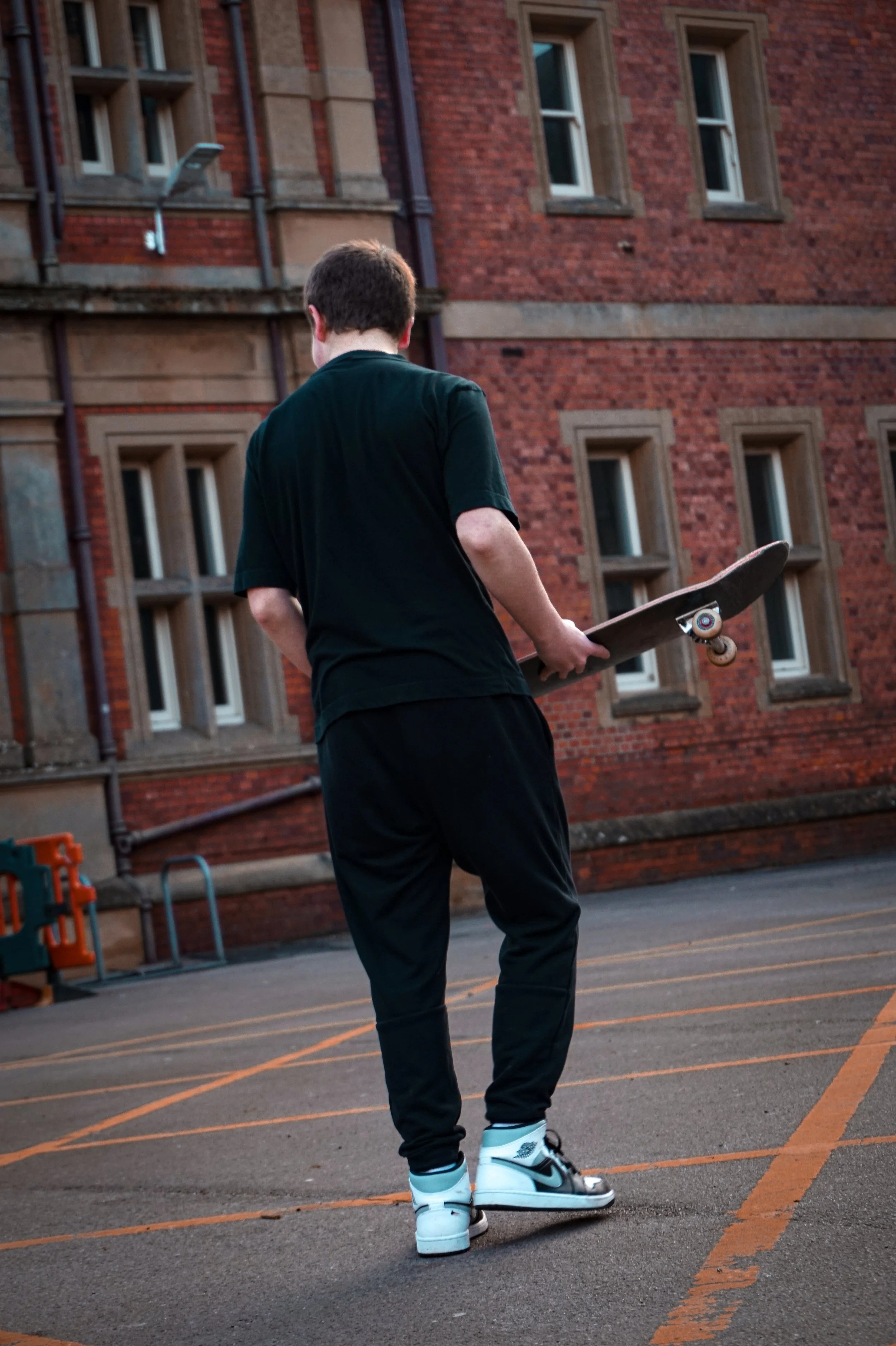 A person holding a skateboard standing on a basketball court with brick school building in the background.