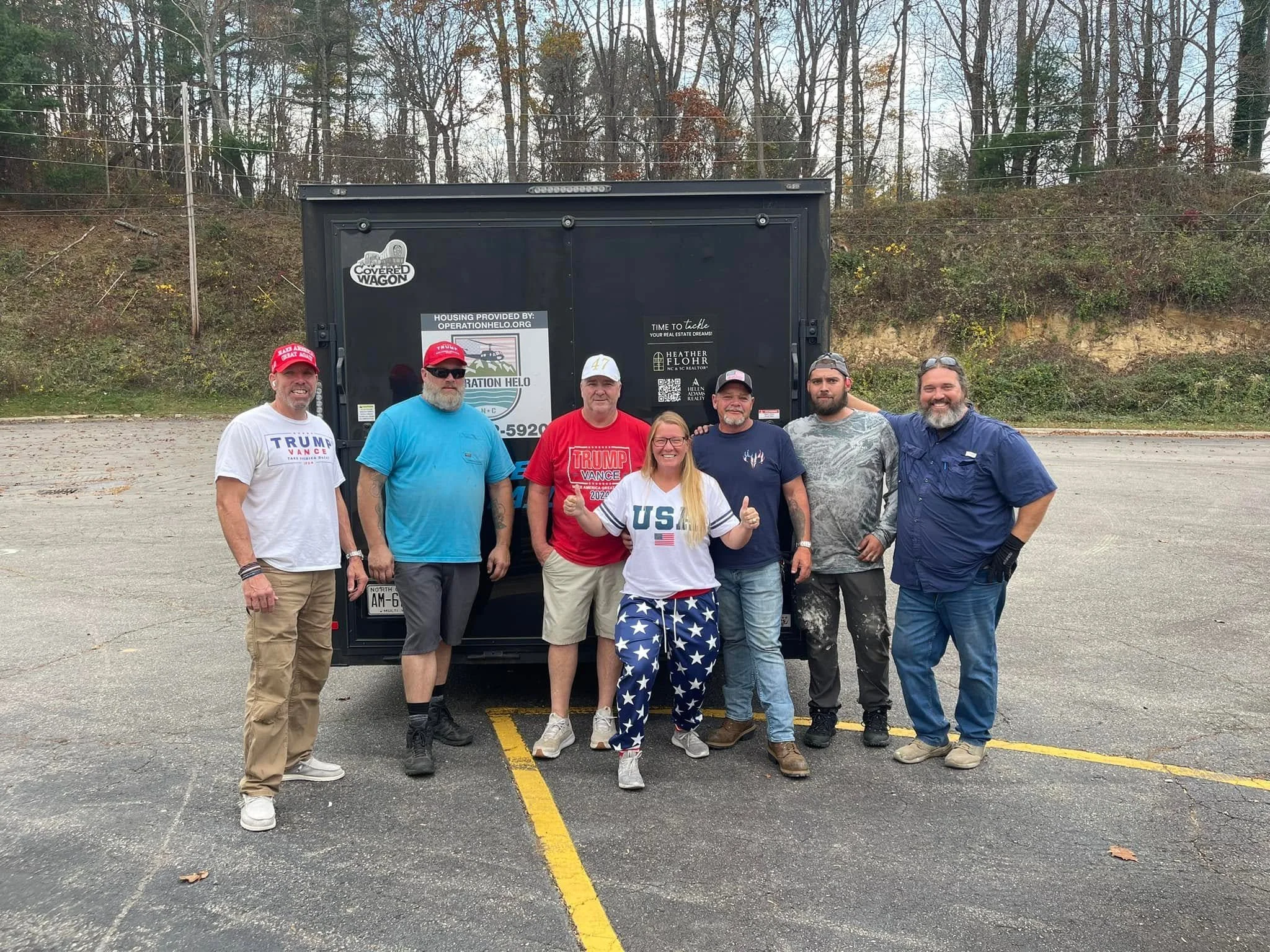 A group of nine people standing in front of a black utility trailer, outdoors on a cleared parking lot with trees in the background. They are smiling and wearing casual clothing, some with political caps and shirts supporting Donald Trump.