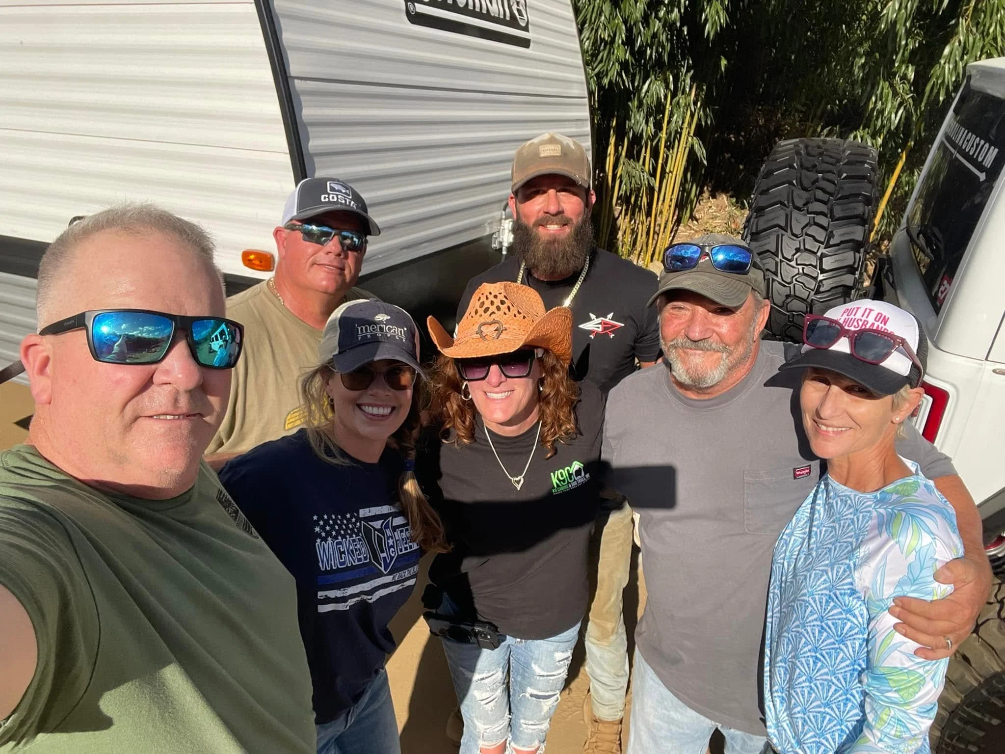 A group of people smiling and taking a selfie outdoors, with a trailer and green foliage in the background.