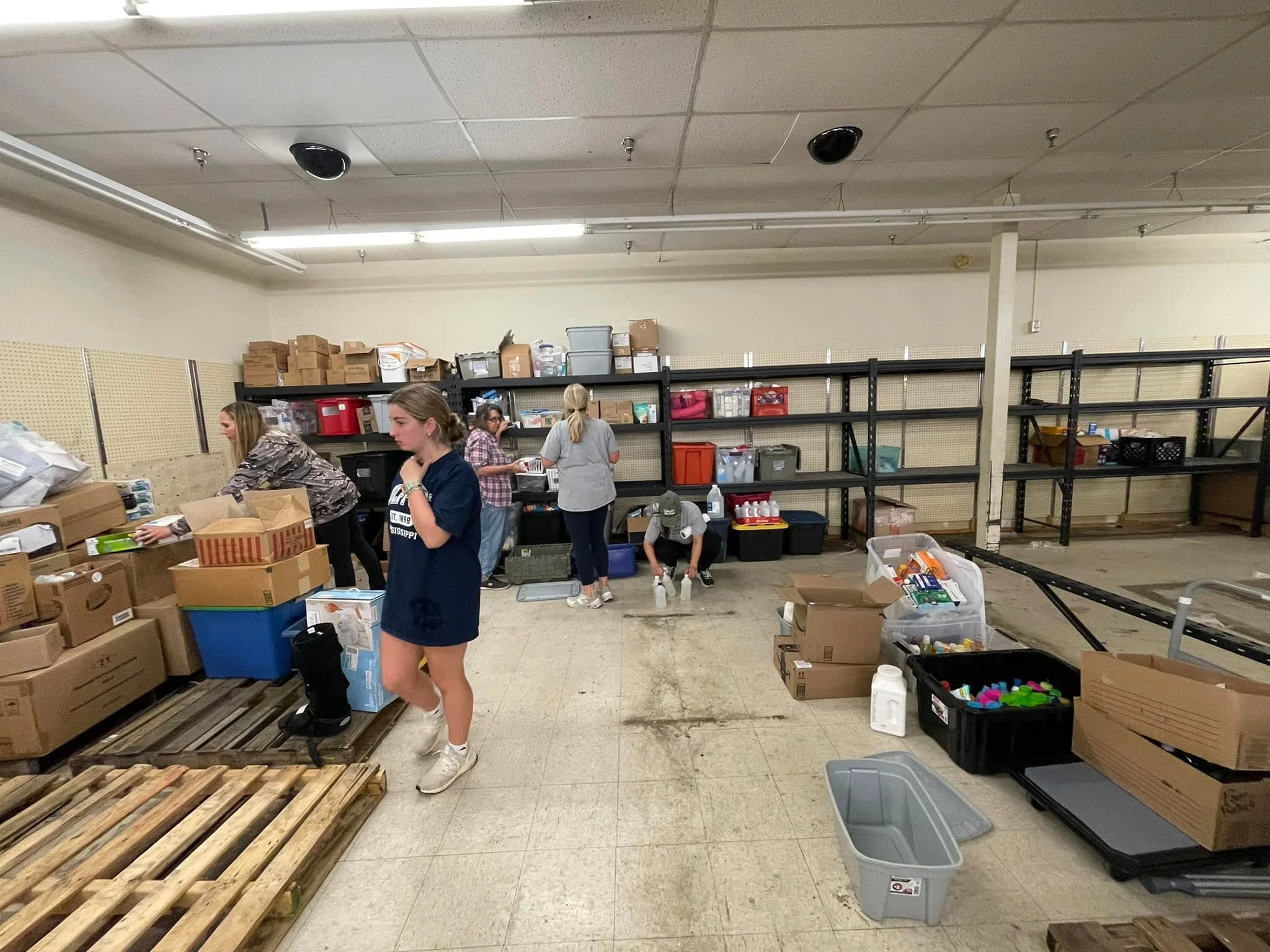 Several people working in a storage room filled with cardboard boxes, plastic containers, and shelving units.