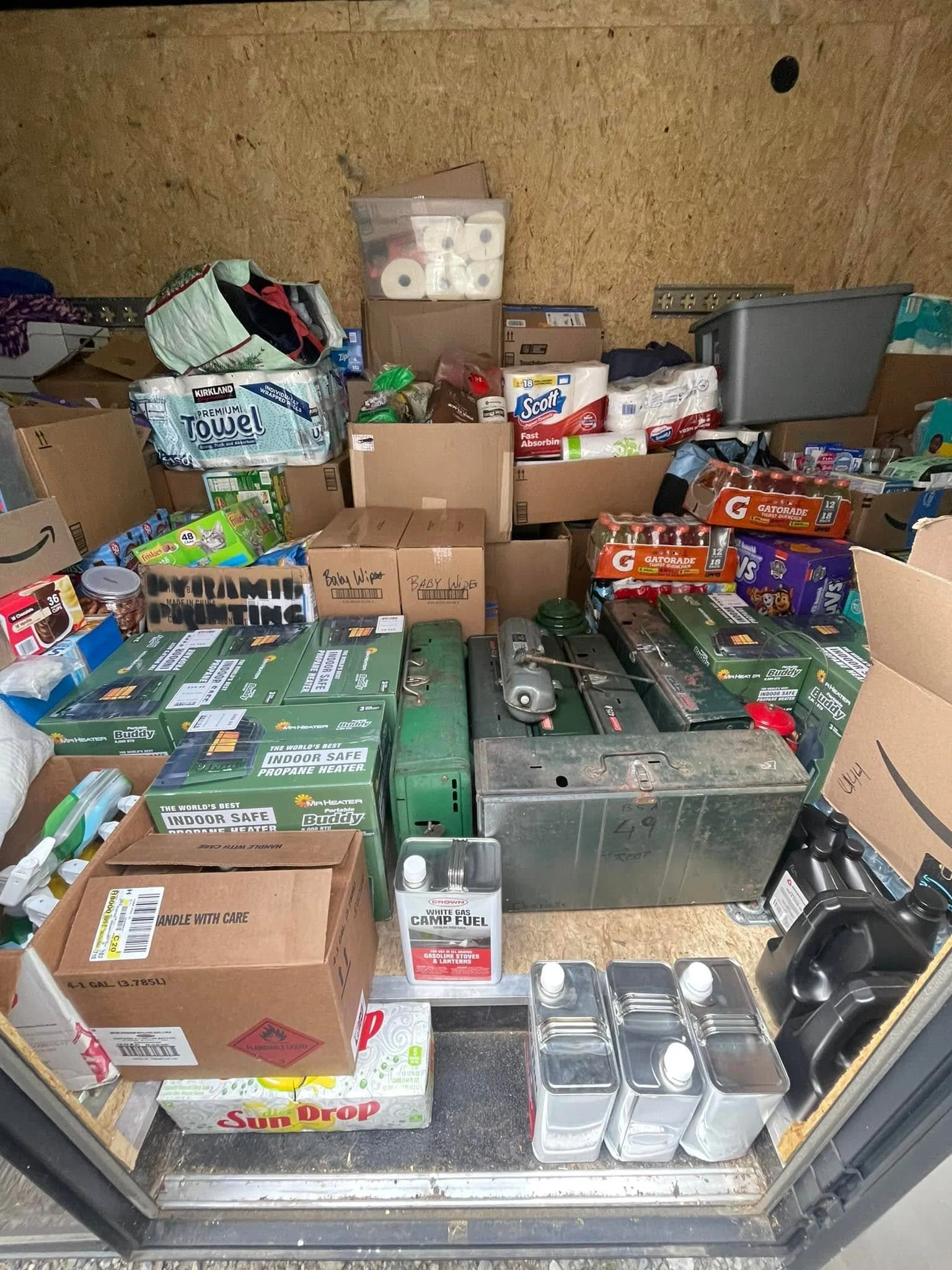 Storage unit filled with boxes and supplies including paper towels, cleaning products, bottled water, and other household items.