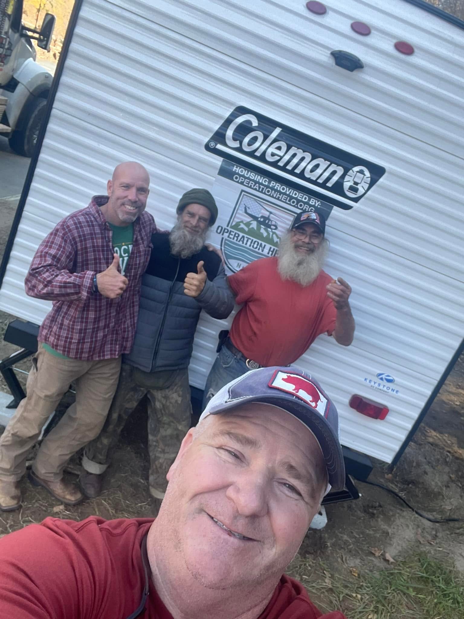A selfie of four men outdoors near a white trailer with the words "Coleman" and 'Operation Helo' logo. The man in the foreground is wearing a red shirt and a cap with a red bear logo. Three men are standing in the background, one giving a thumbs up, 