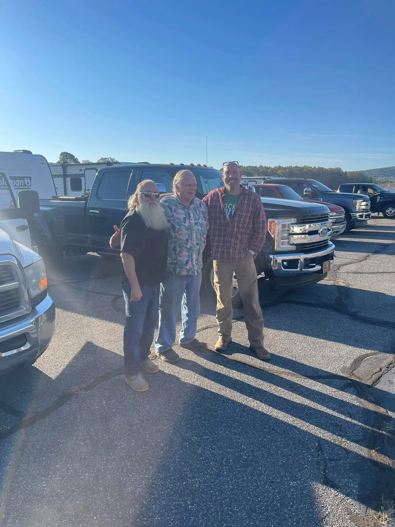 Three men standing in a parking lot next to a row of trucks and SUVs, smiling and posing for the camera, with clear blue sky and distant trees in the background.