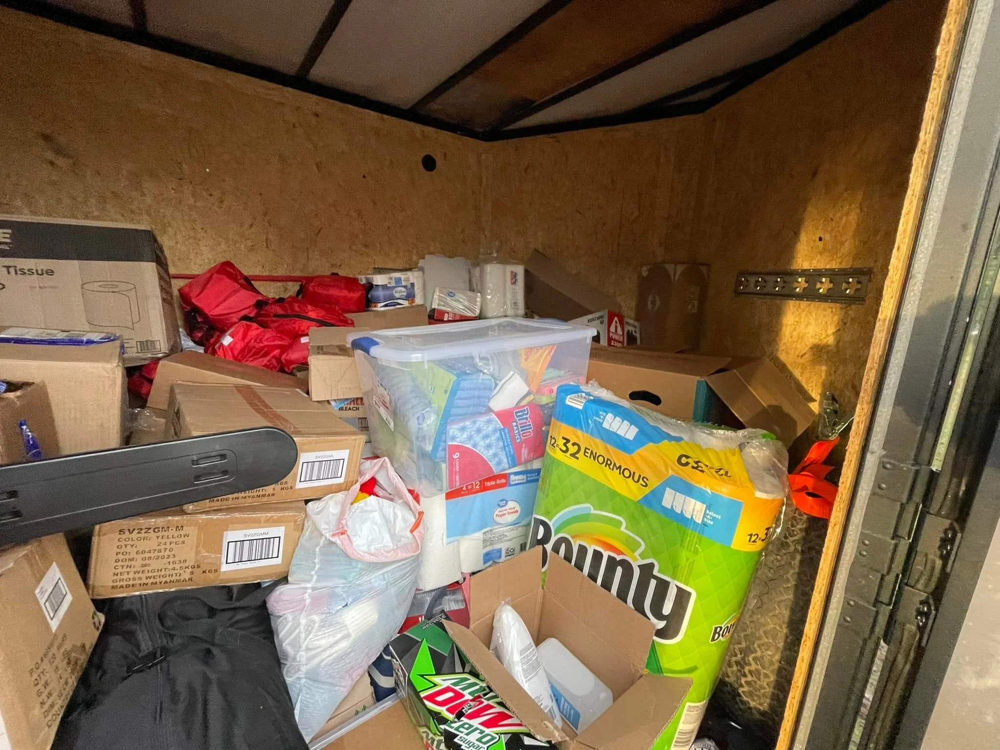 A cluttered storage space or closet filled with various boxes, packages, and household items, with a wood-paneled wall and a yellow-flowered wallpapered surface.