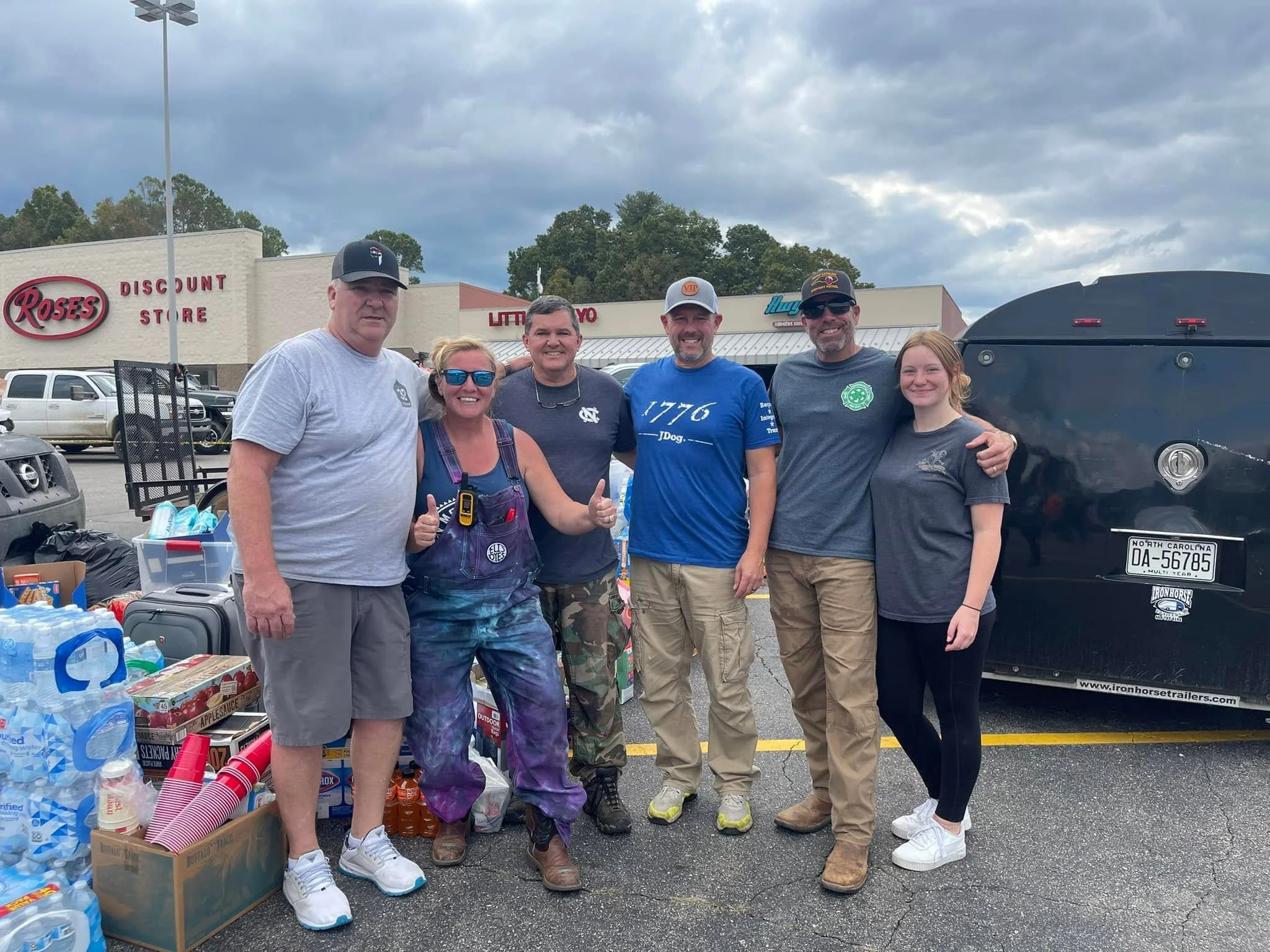 Group of six people standing in a parking lot in front of a building, smiling, with donation items like bottled water and cups around them.