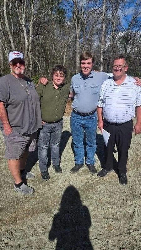 Four men standing outdoors on a sunny day, smiling at the camera, with leafless trees and a blue sky in the background.
